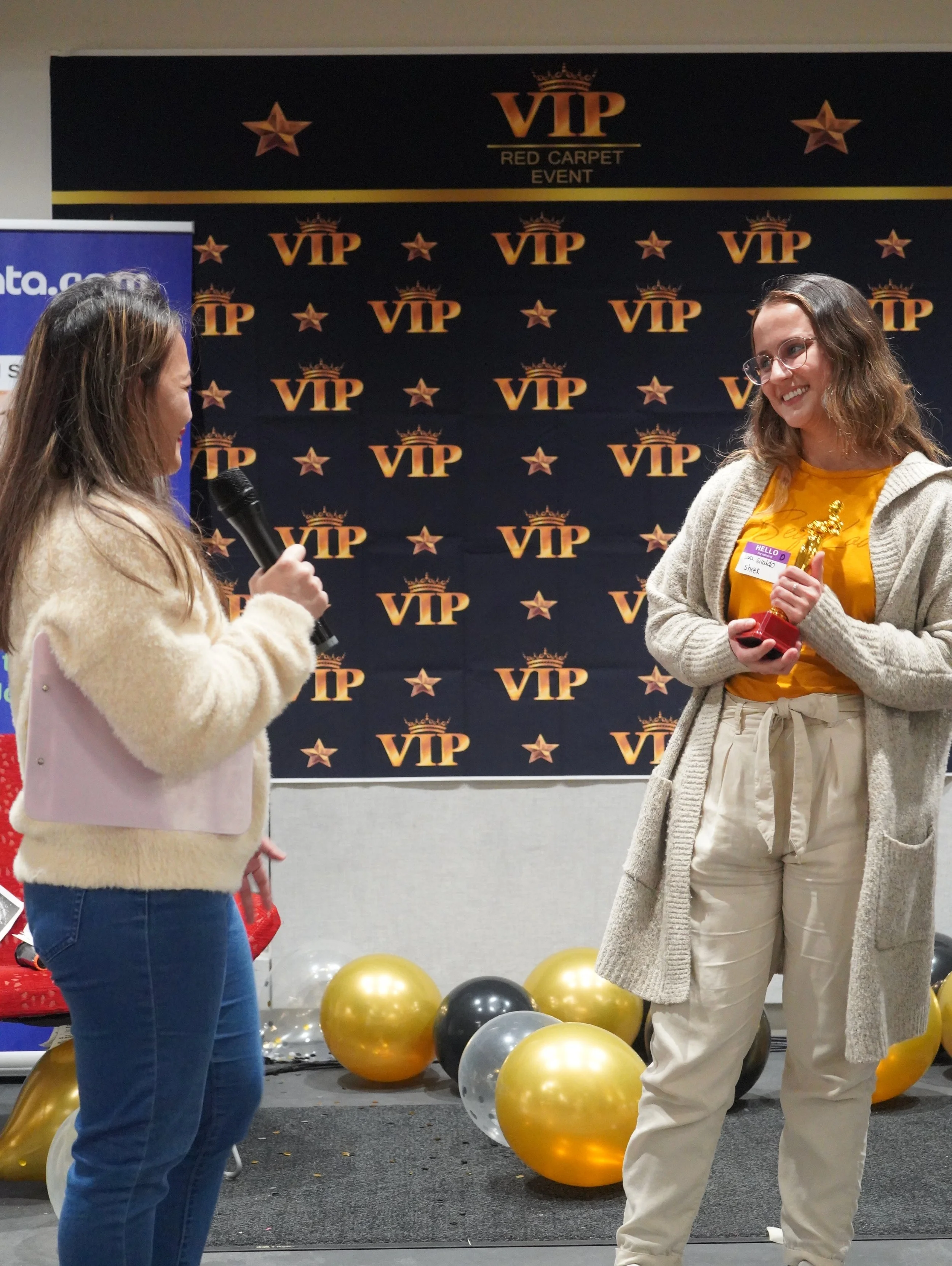 Two women speaking at an event with a VIP-themed backdrop decorated with gold and black balloons, stars, and balloons.