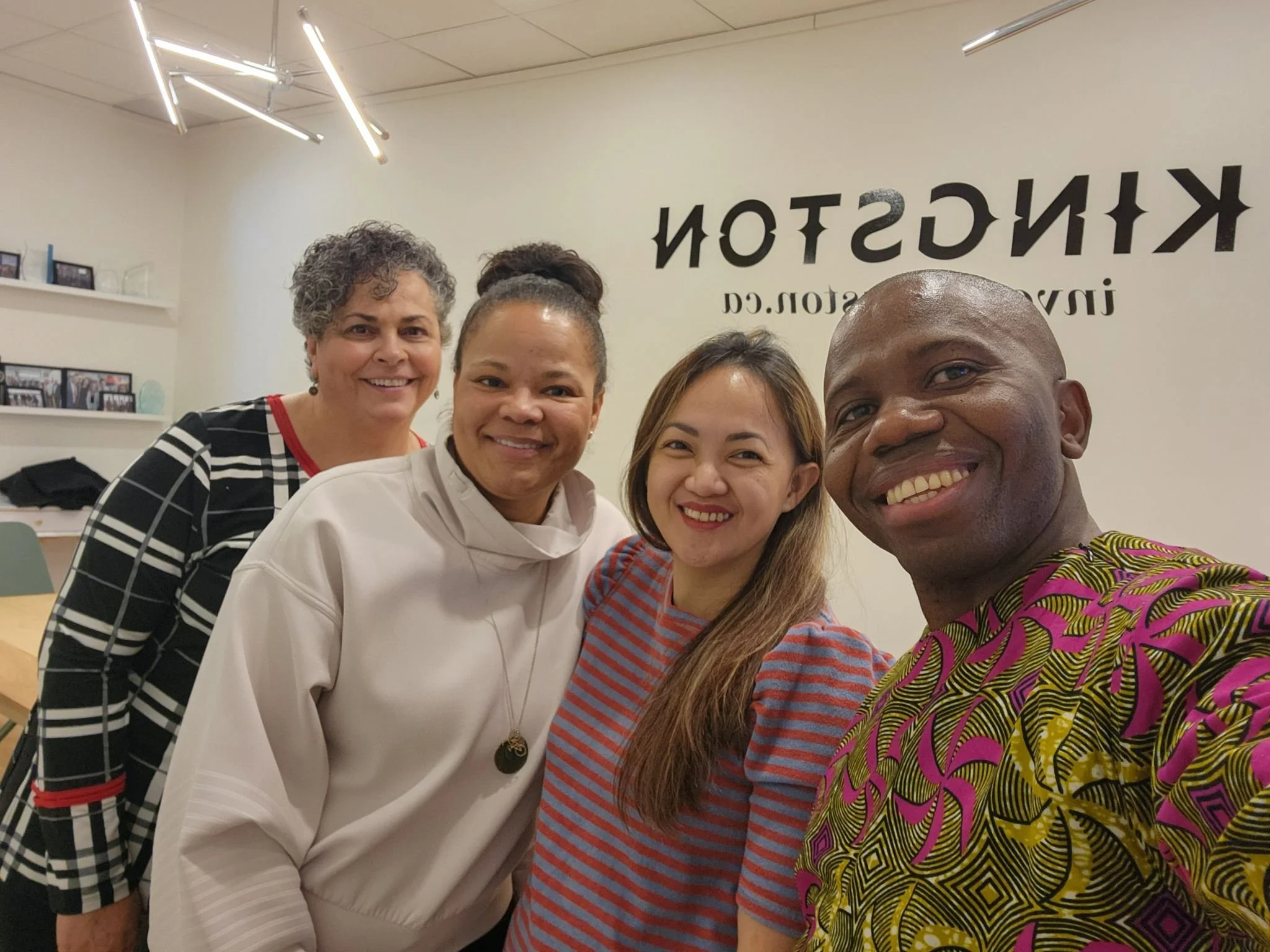 Four diverse women smiling and taking a selfie together in an office or community space, with a sign reading 'KINGSTON' behind them.