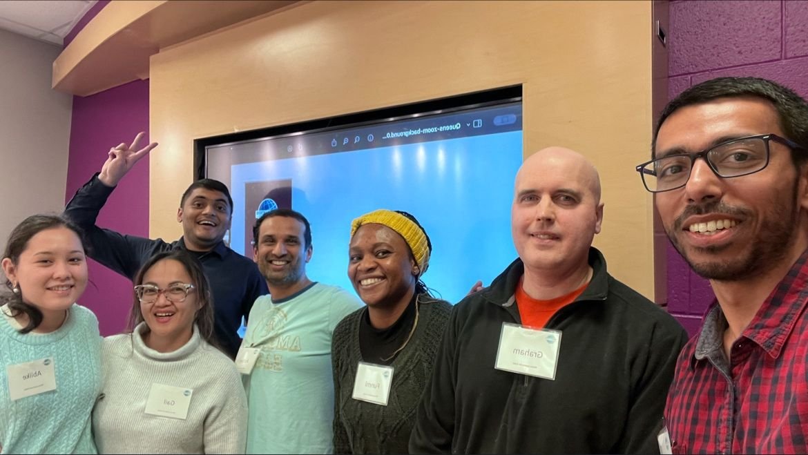 Group of seven people smiling for a photo in a room with a large screen behind them.