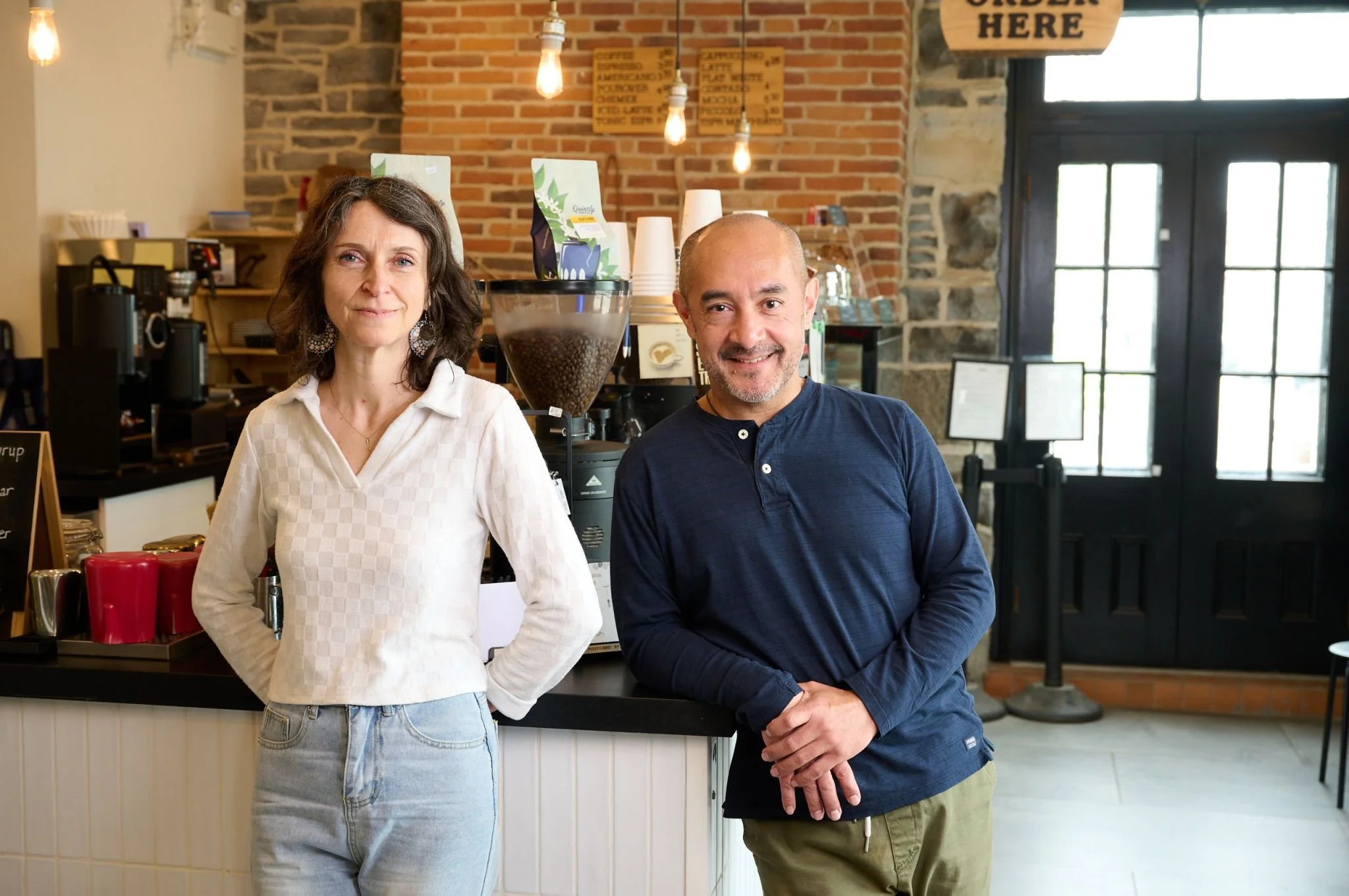 A man and woman standing inside a coffee shop near the counter, smiling at the camera. The woman has shoulder-length brown hair, wearing a white checked shirt, and the man is bald, wearing a navy blue long-sleeve shirt. The background shows coffee brewing equipment, a sign that says "Order Here," and a large window with natural light.