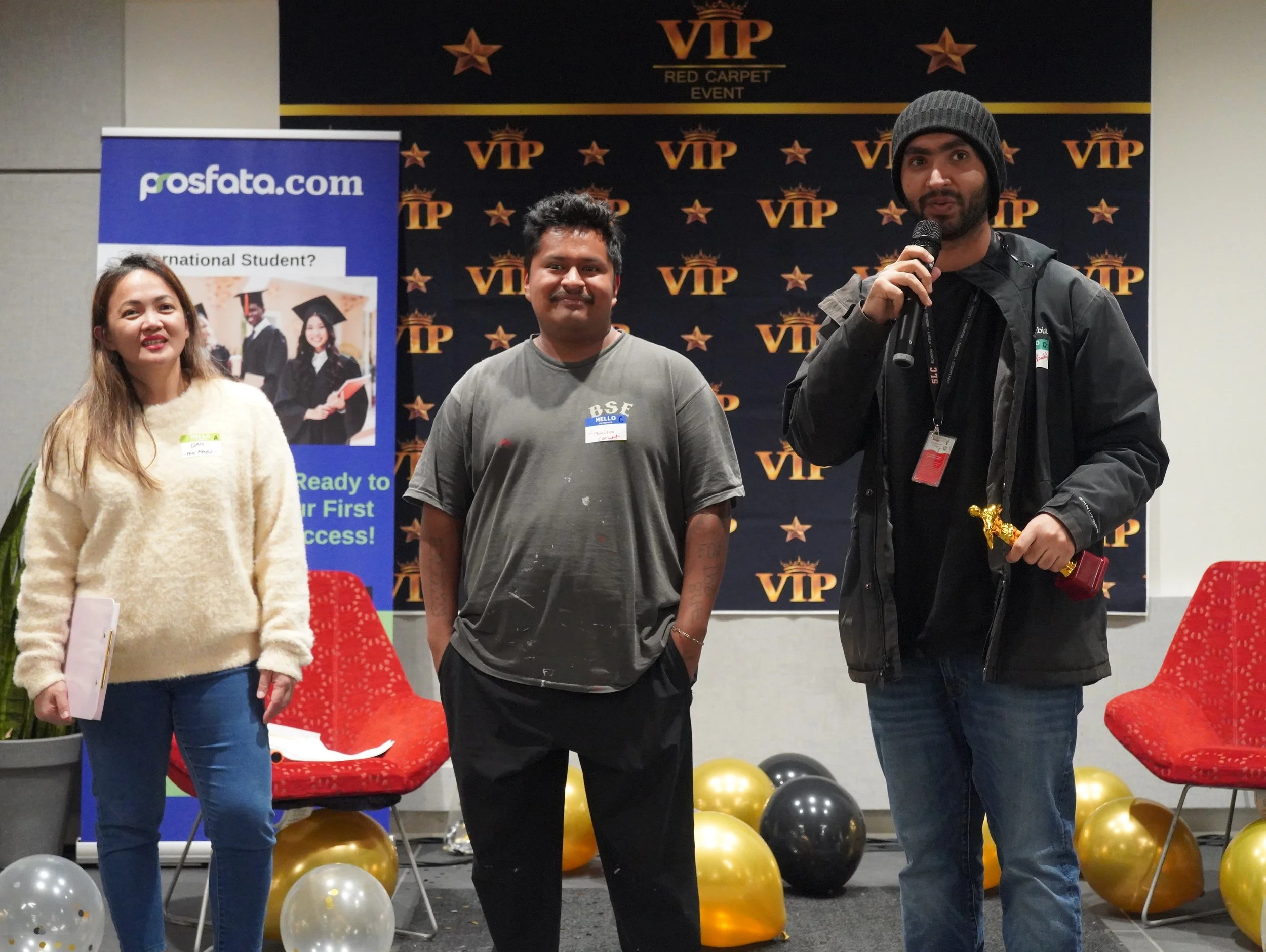 Three people standing on a stage at an event, with a VIP backdrop and balloons on the floor. The person on the right is speaking into a microphone, holding a small trophy, and wearing a beanie. The woman on the left is holding a folder, and the man in the middle is smiling with his hands in his pockets.