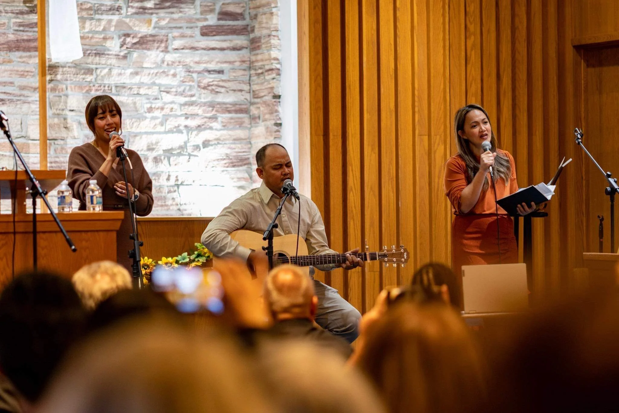 Three performers on stage; two women singing into microphones and a man playing an acoustic guitar. Audience members are visible in the foreground.
