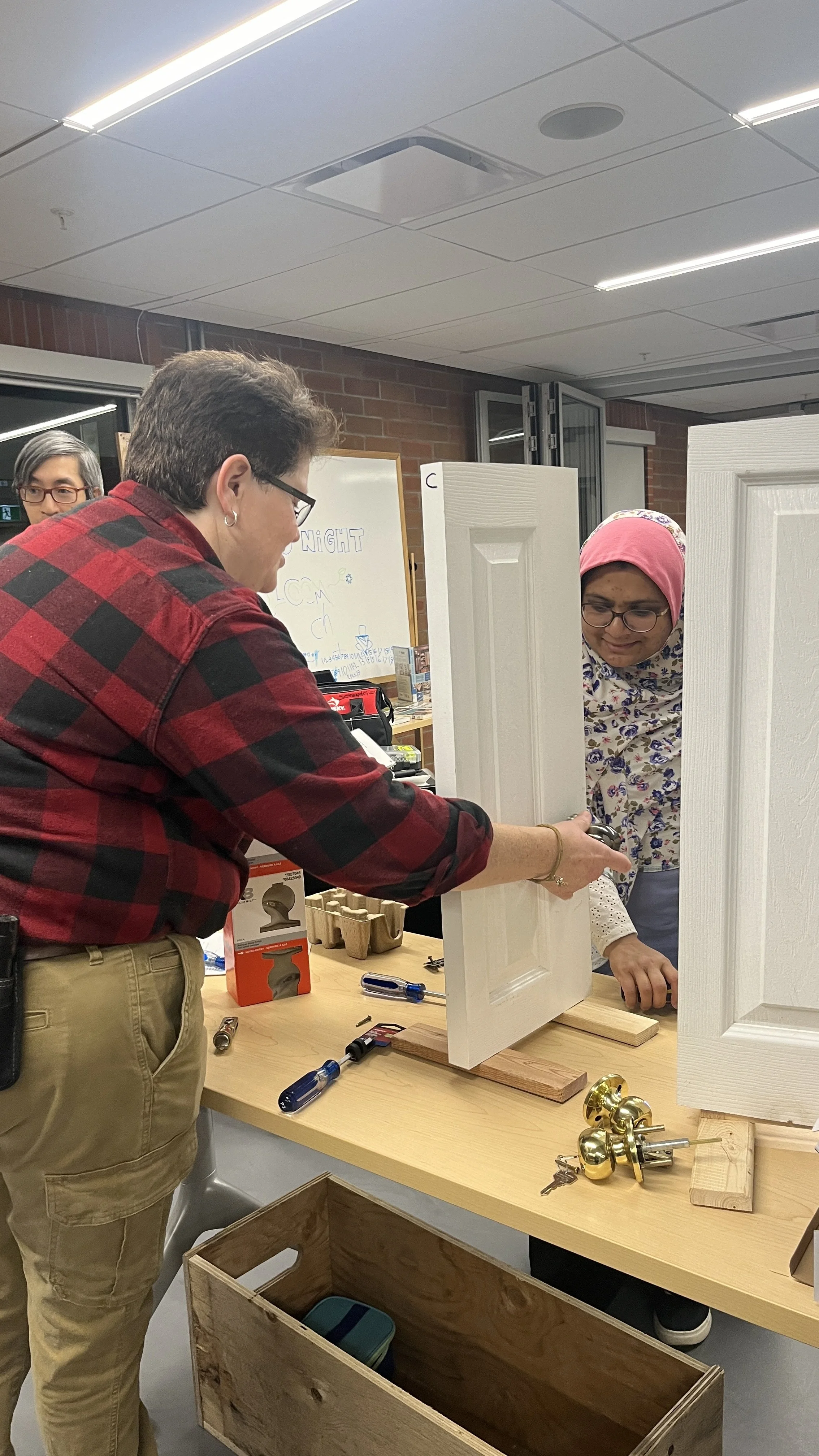A woman with a pink headscarf and glasses looks concerned as another woman demonstrates a door lock on a white door panel at a workshop or class, with tools and hardware on the table.