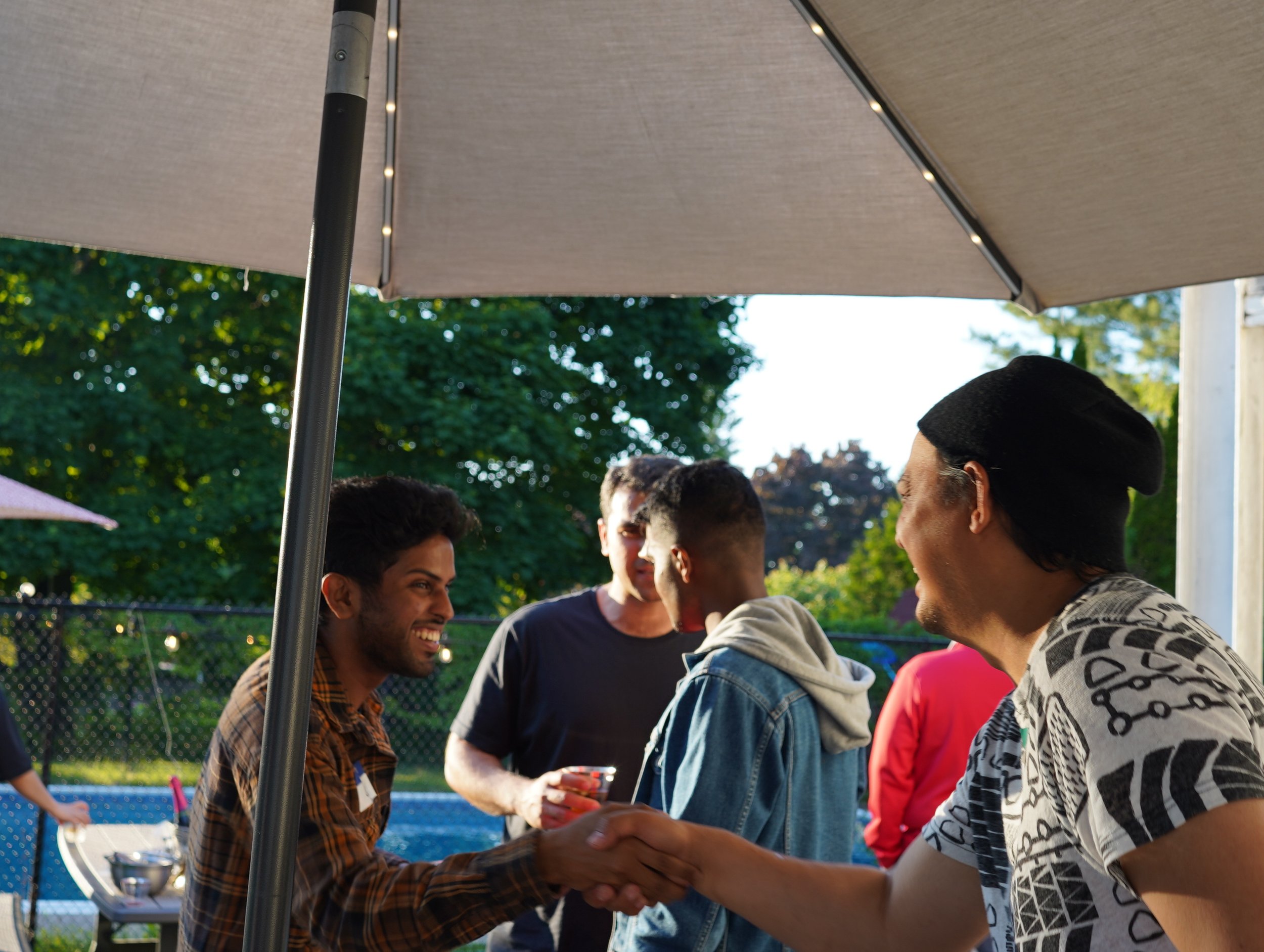 People at a social gathering outdoors under an umbrella, shaking hands, with trees and a fence in the background.