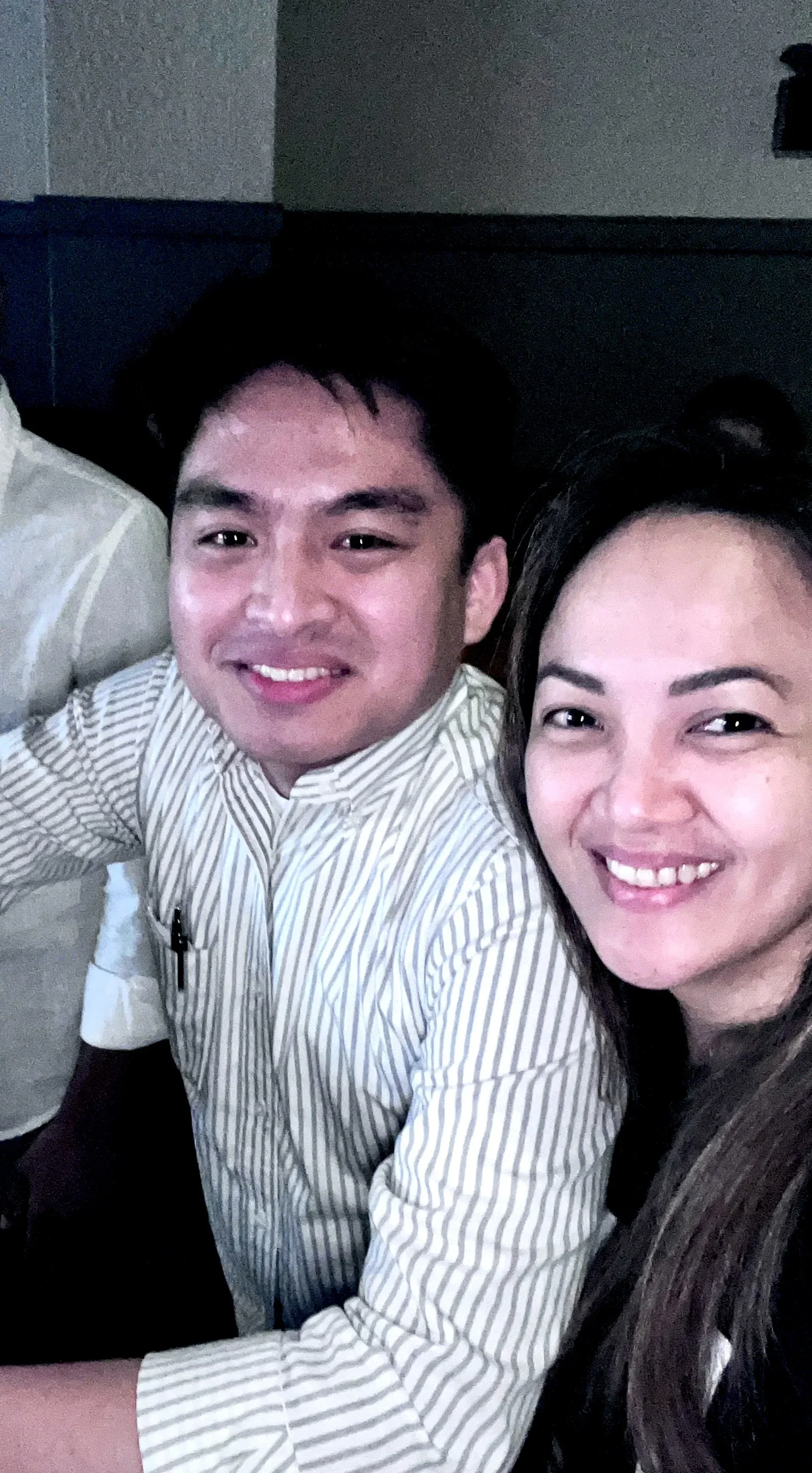 Two smiling people, a man and a woman, sitting close together for a selfie in an indoor setting.