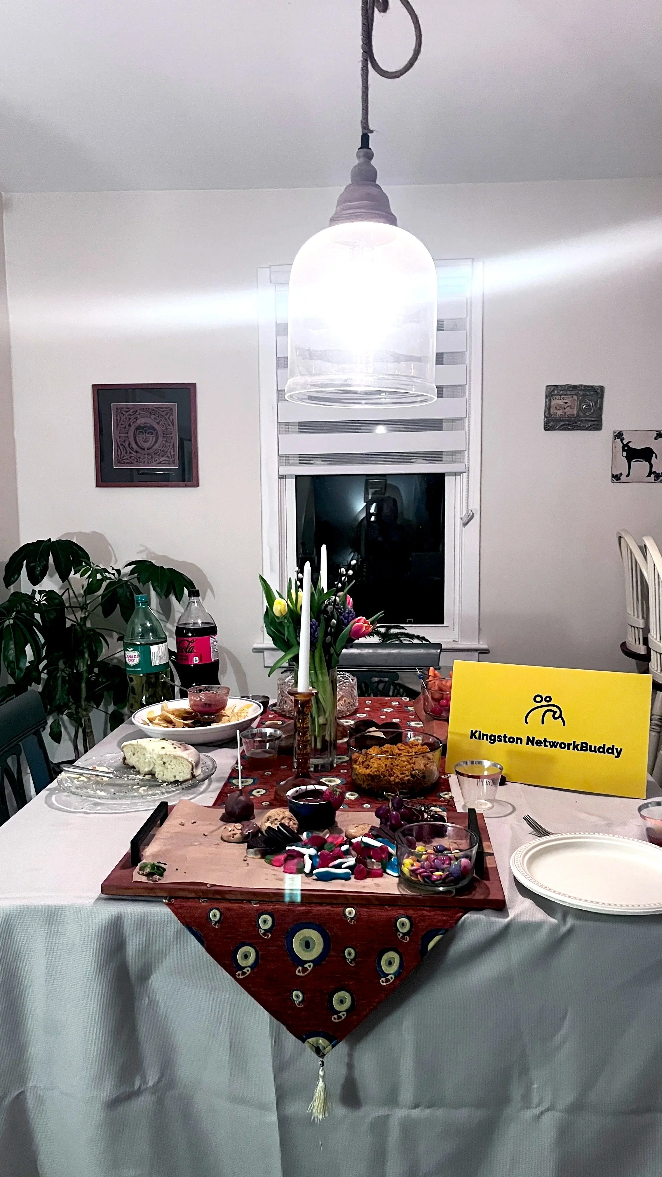 A dining table set for a celebration with various foods, desserts, drinks, a yellow sign that says 'Kingston NetworkBuddy', a vase with tulips, and a chandelier hanging above.