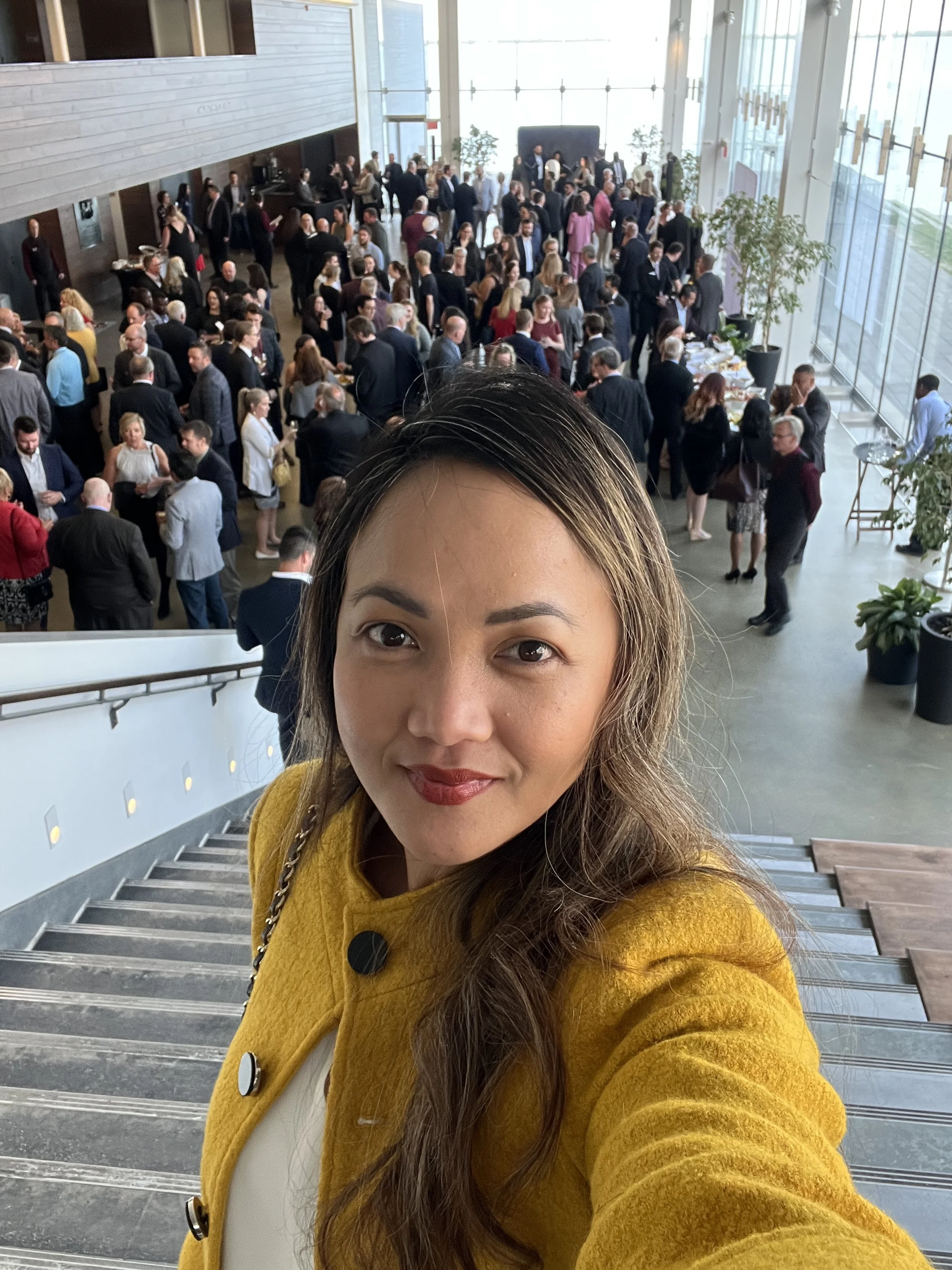 A woman taking a selfie on a staircase at a crowded indoor event with many people gathered in the background, some near a buffet table and large windows.