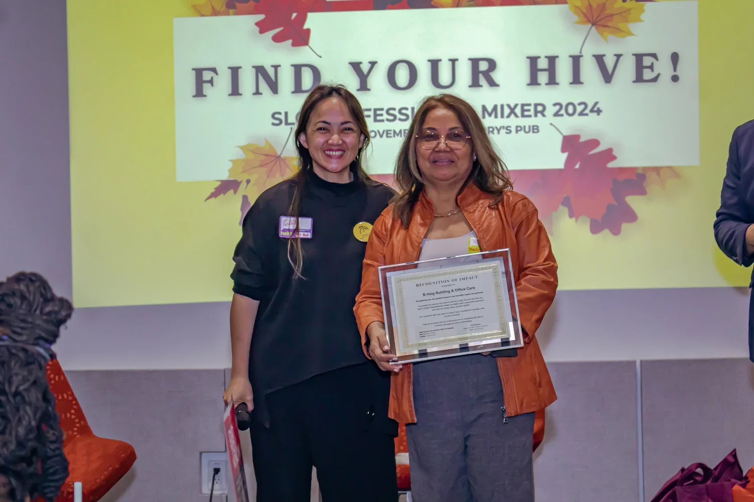 Two women standing in front of a presentation screen, one holding a framed certificate and the other smiling, during an event titled 'Find Your Hive!'. The background features autumn leaves and the event details.