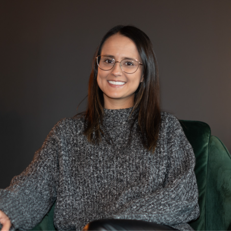 A woman with shoulder-length dark hair, wearing glasses and a grey knitted sweater, smiling while sitting on a green chair against a grey wall background.