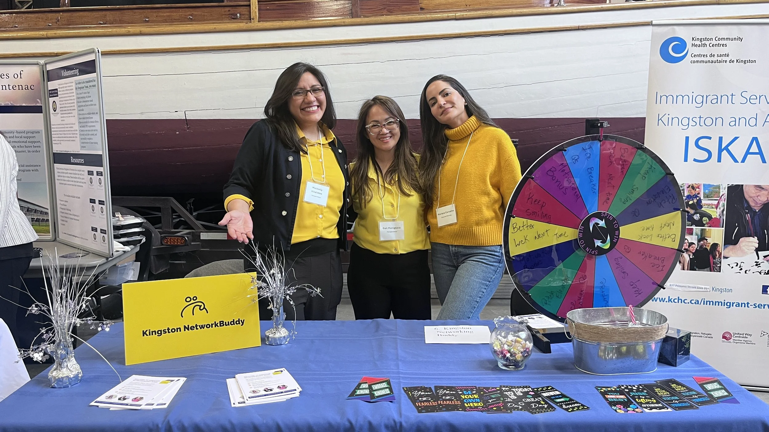 Three women standing behind a table at an event, smiling at the camera. The table displays a sign that says "Kingston NetworkBuddy," pamphlets, and brochures. To the right, a colorful spin wheel is set up next to a large banner that reads "Kingston Community Health Centres" and "Immigrant Services Kingston and Area." The women are wearing yellow shirts, and one is wearing a black jacket. The setting appears to be indoors, possibly at a health or community event.