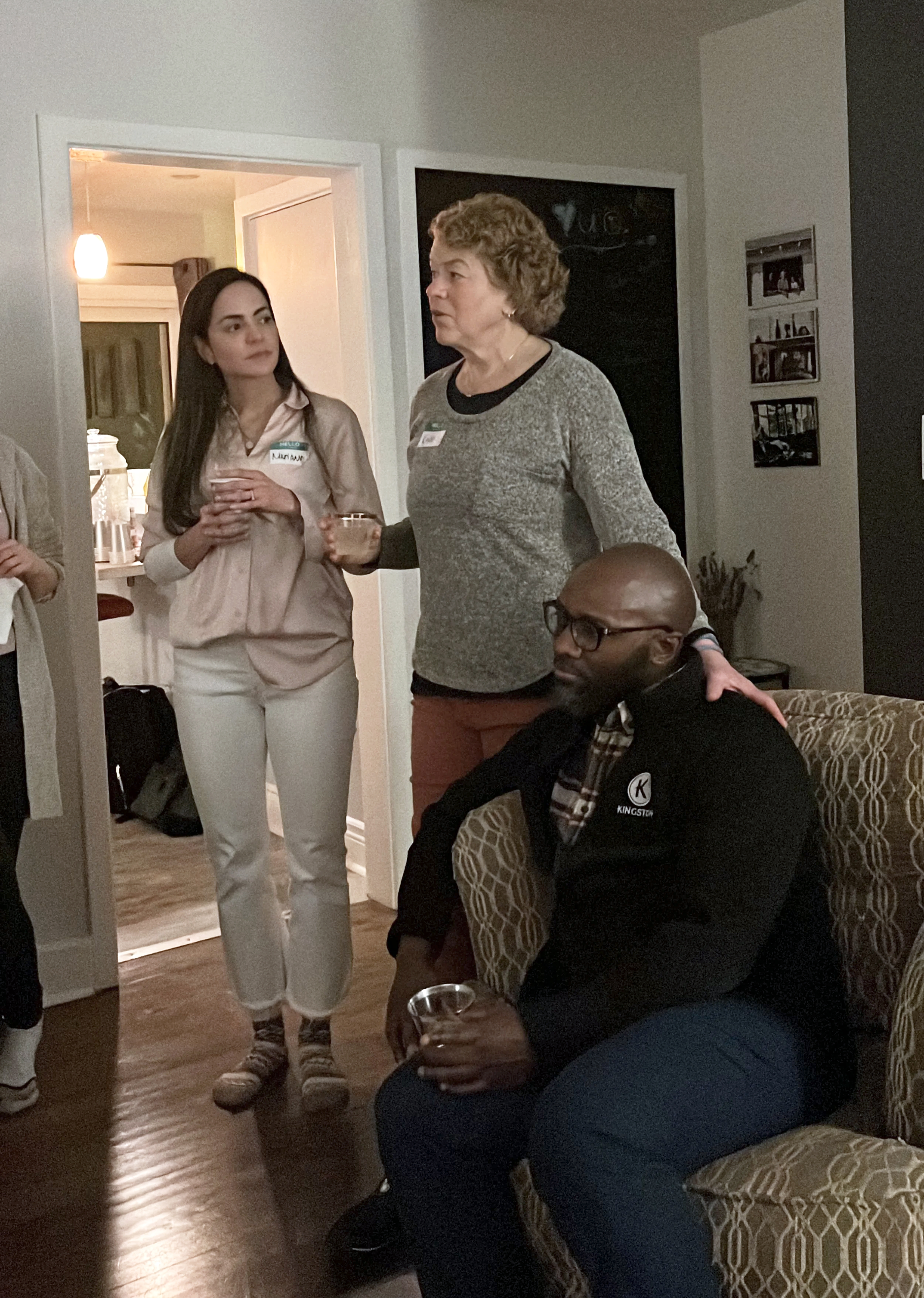 People socializing in a living room at a gathering, with three individuals visible: a woman with curly hair in a gray sweater holding a drink, a young woman with dark hair holding a cup, and a man with glasses sitting on a patterned sofa holding a drink. There is a doorway leading to a kitchen area in the background.