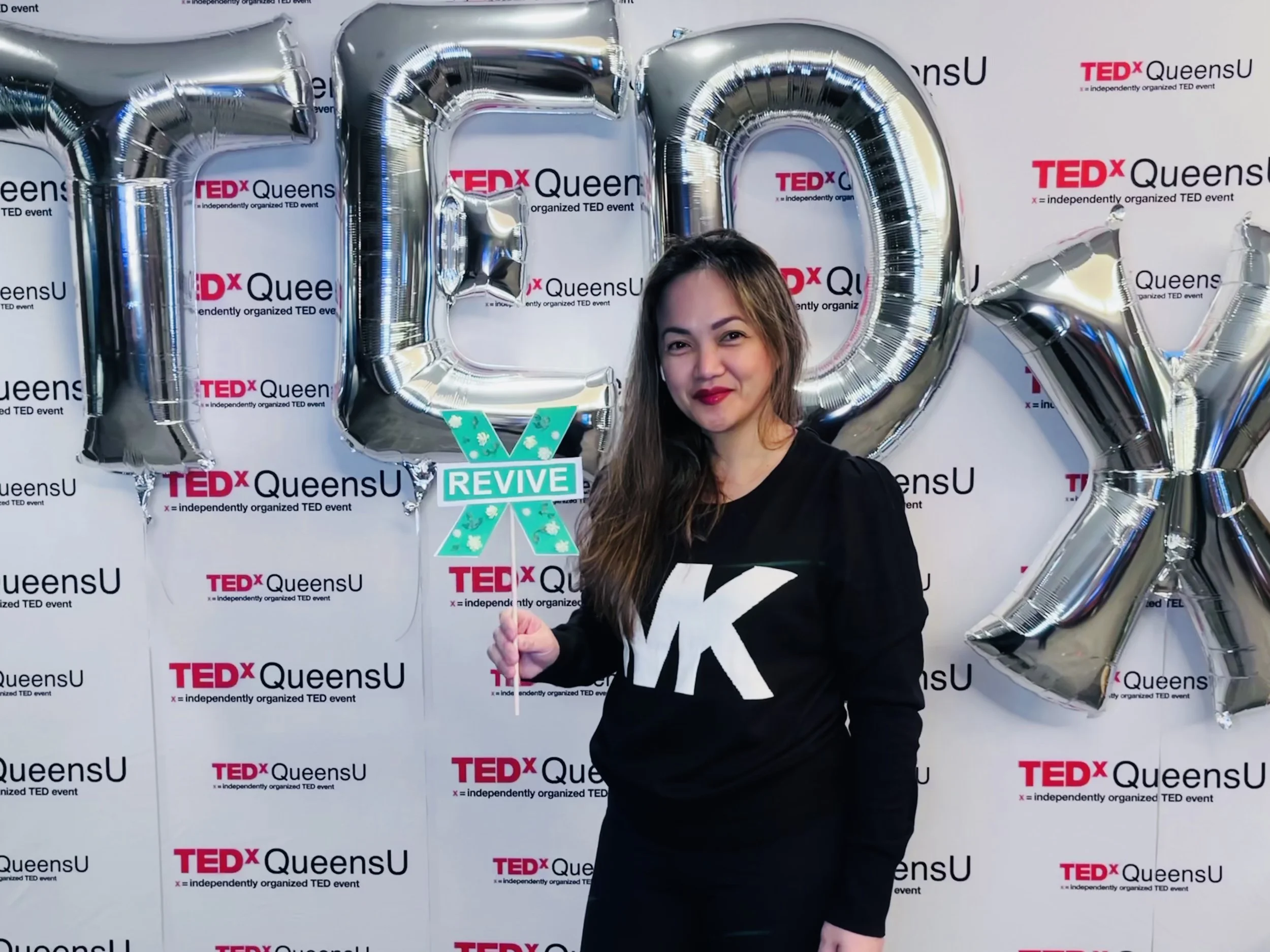 Woman standing in front of TEDx QueensU backdrop with large balloon letters spelling 'LOVE' and holding a sign that says 'REVIVE.'