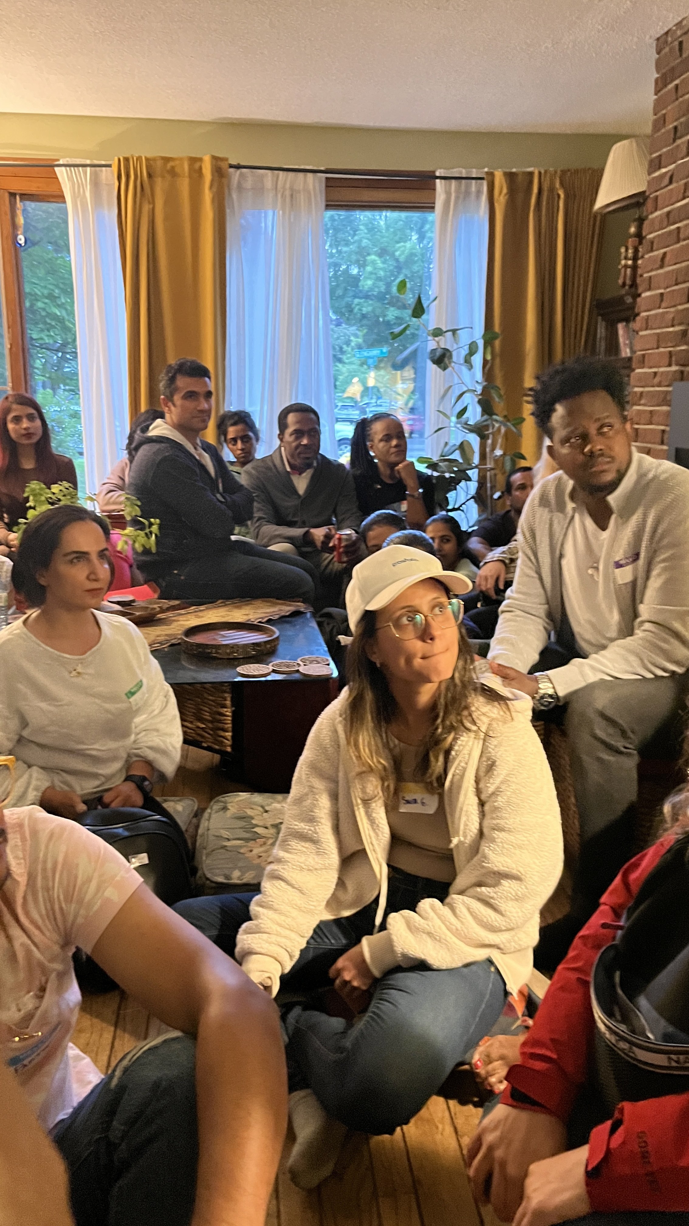 Group of people seated indoors, some sitting on the floor and others on a piece of furniture, in a cozy room with large windows, yellow and white curtains, and a brick fireplace.