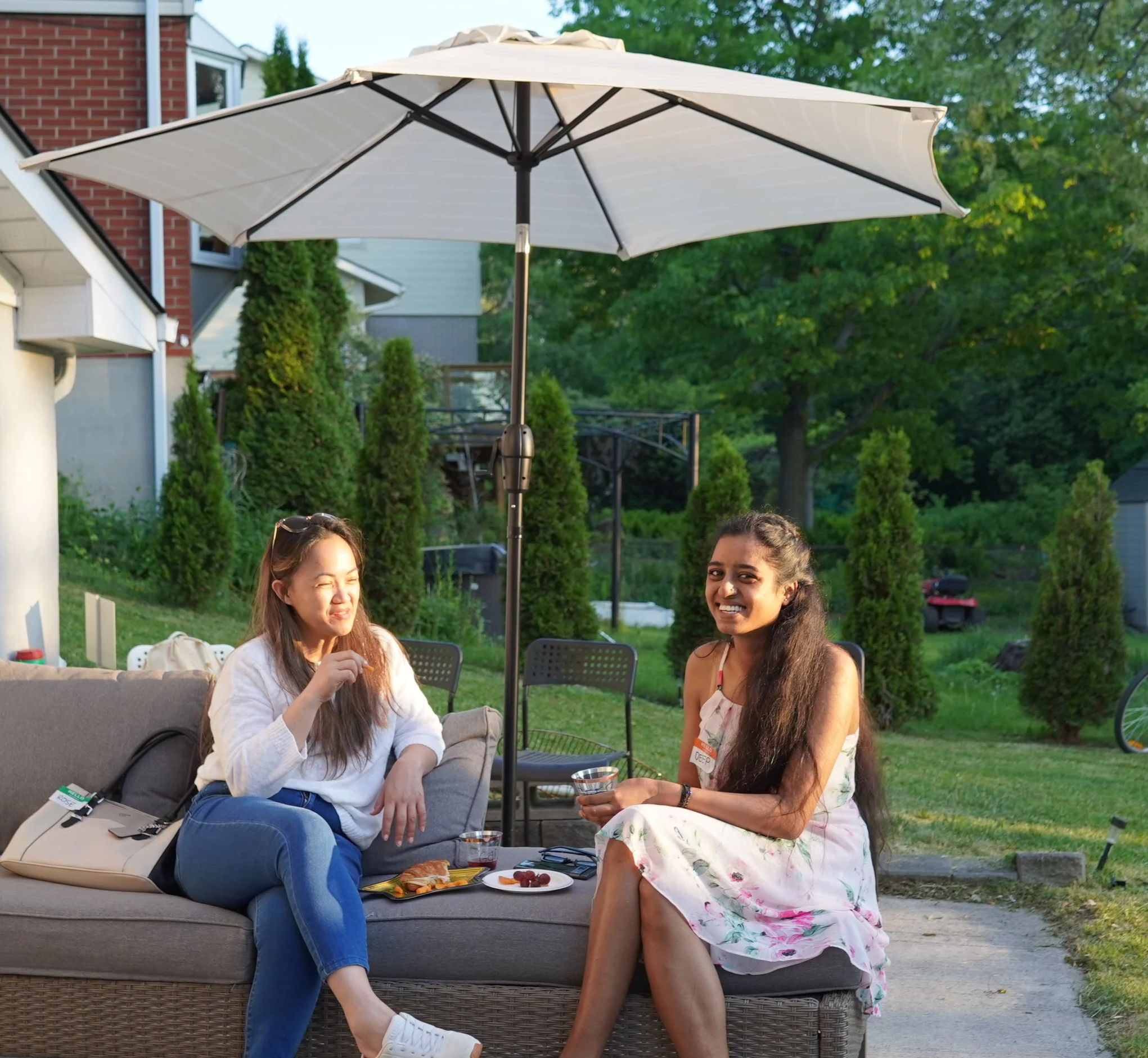 Two women sitting outside on a couch under a large patio umbrella, enjoying drinks and food in a backyard with green trees and houses in the background.