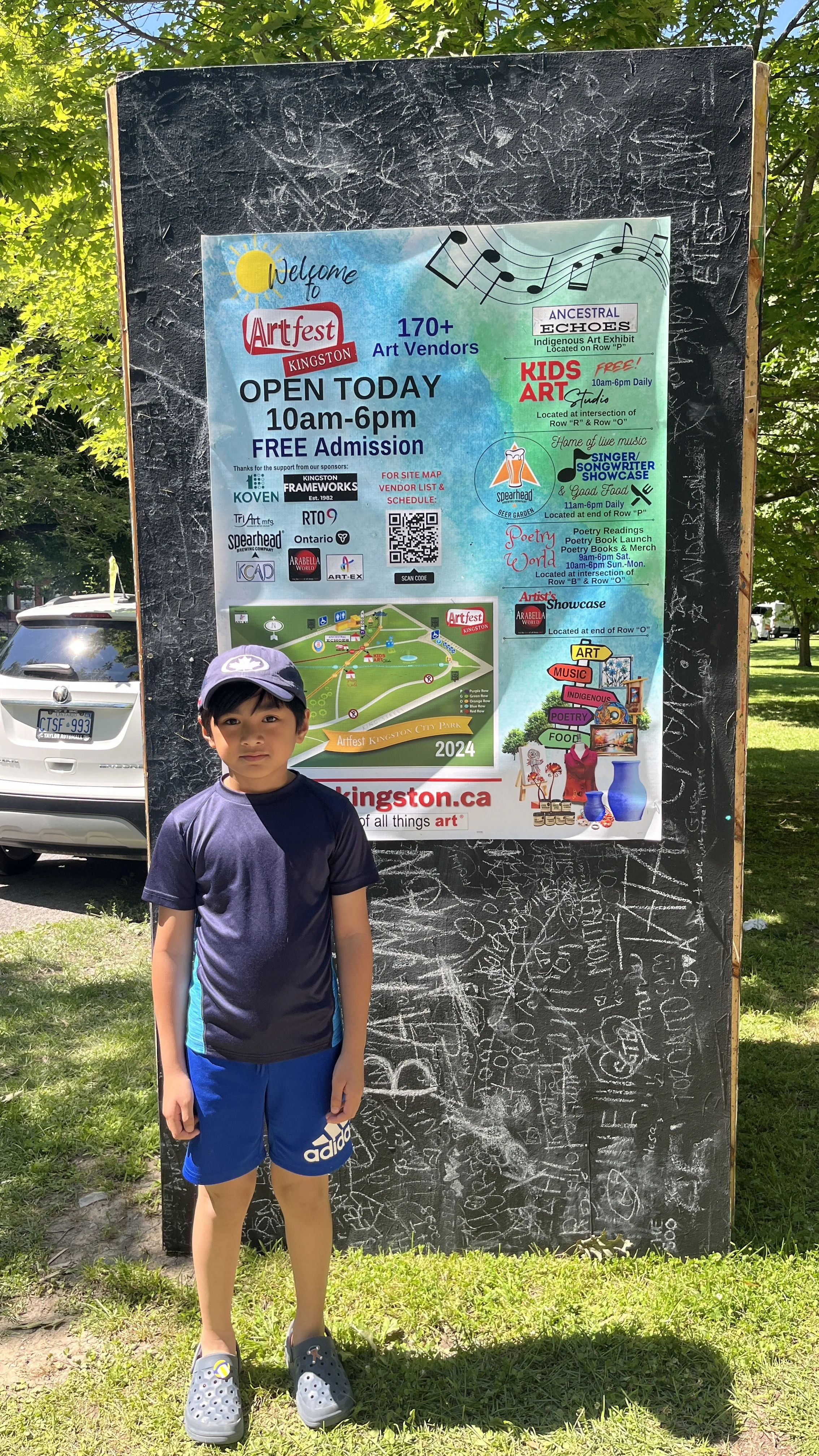 A young boy standing in front of a large blackboard with art festival information, including details about vendors, art exhibits, kid's art, poetry readings, and scheduled activities at Artfest Kingston.