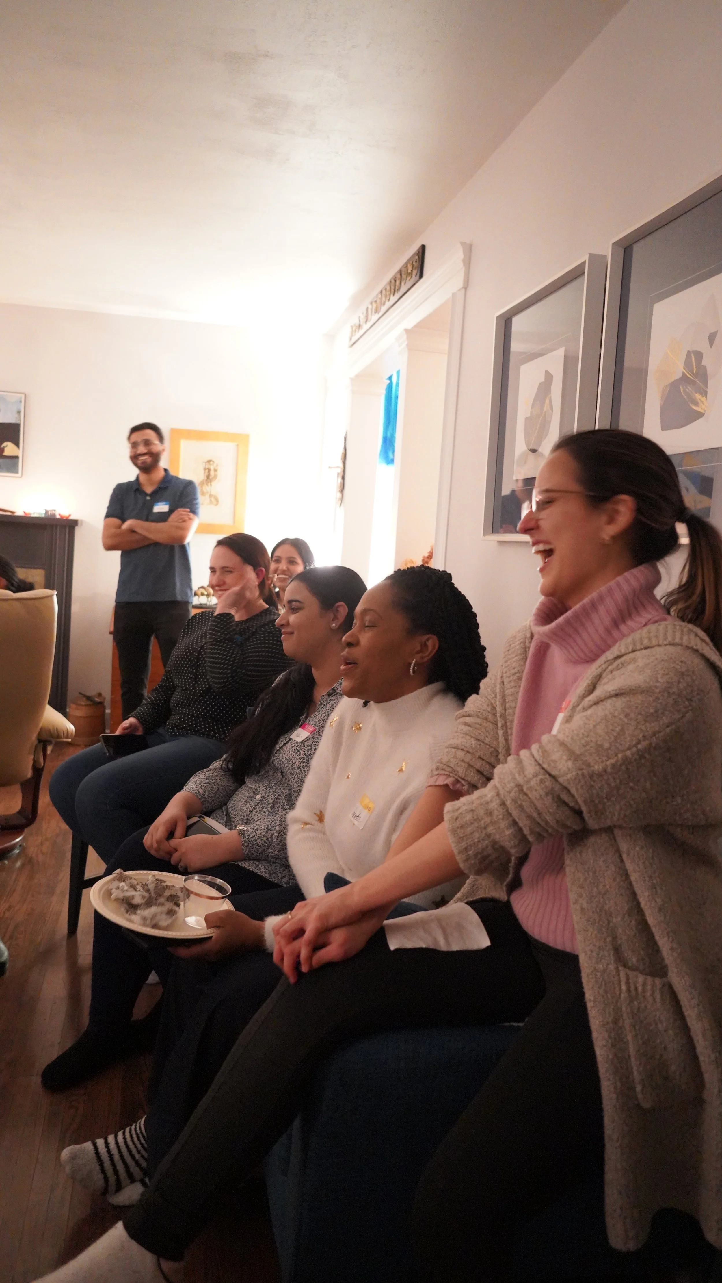 Women sitting in a row, laughing, with a man standing in the background, smiling and crossing his arms, in a cozy indoor setting.