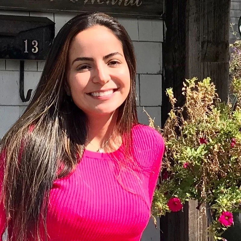 A smiling young woman with long brown hair wearing a bright pink top, standing outdoors next to a bush with pink flowers.