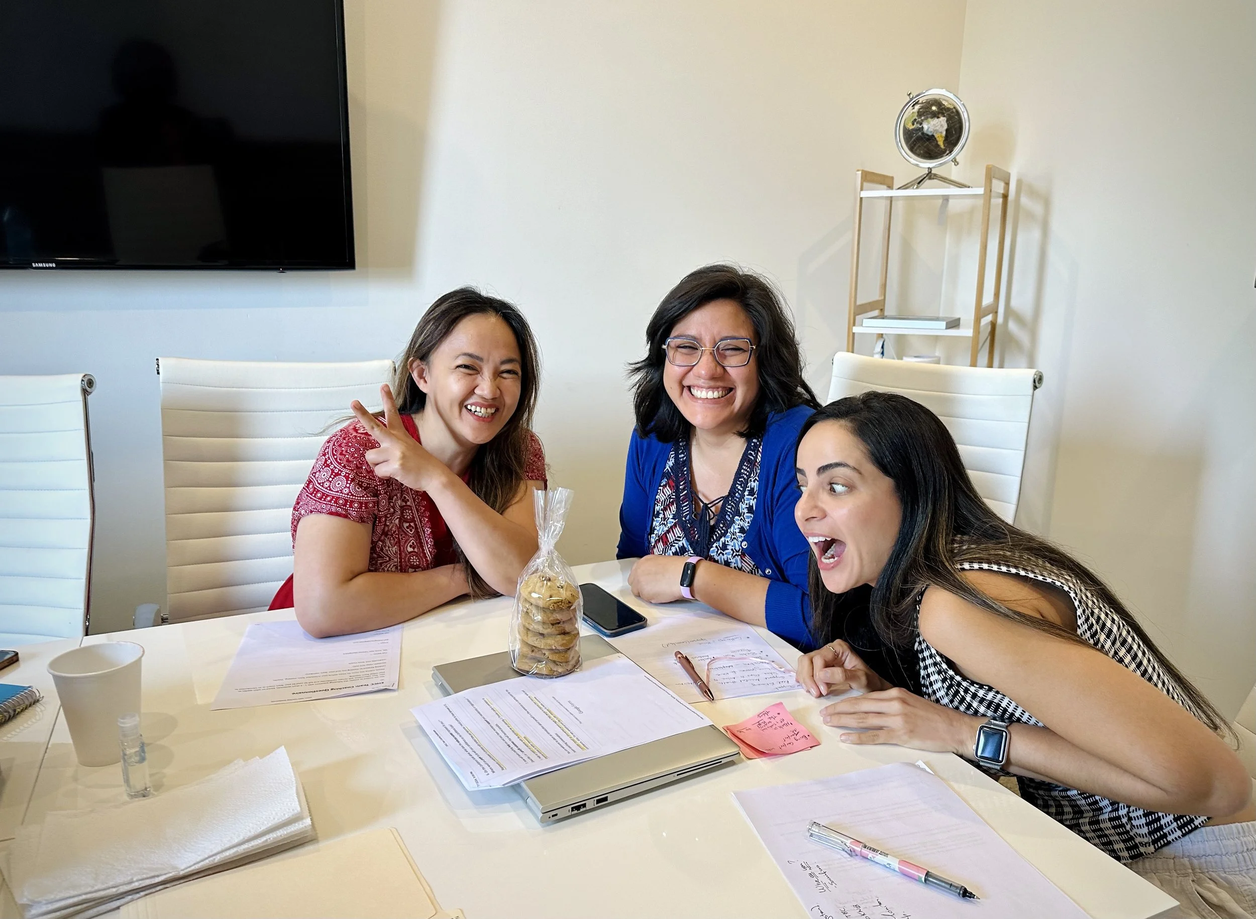 Three women sitting around a conference table, smiling and laughing, with papers, notebooks, pens, and a laptop in front of them, in a bright room with a television on the wall and a shelf with a globe in the background.