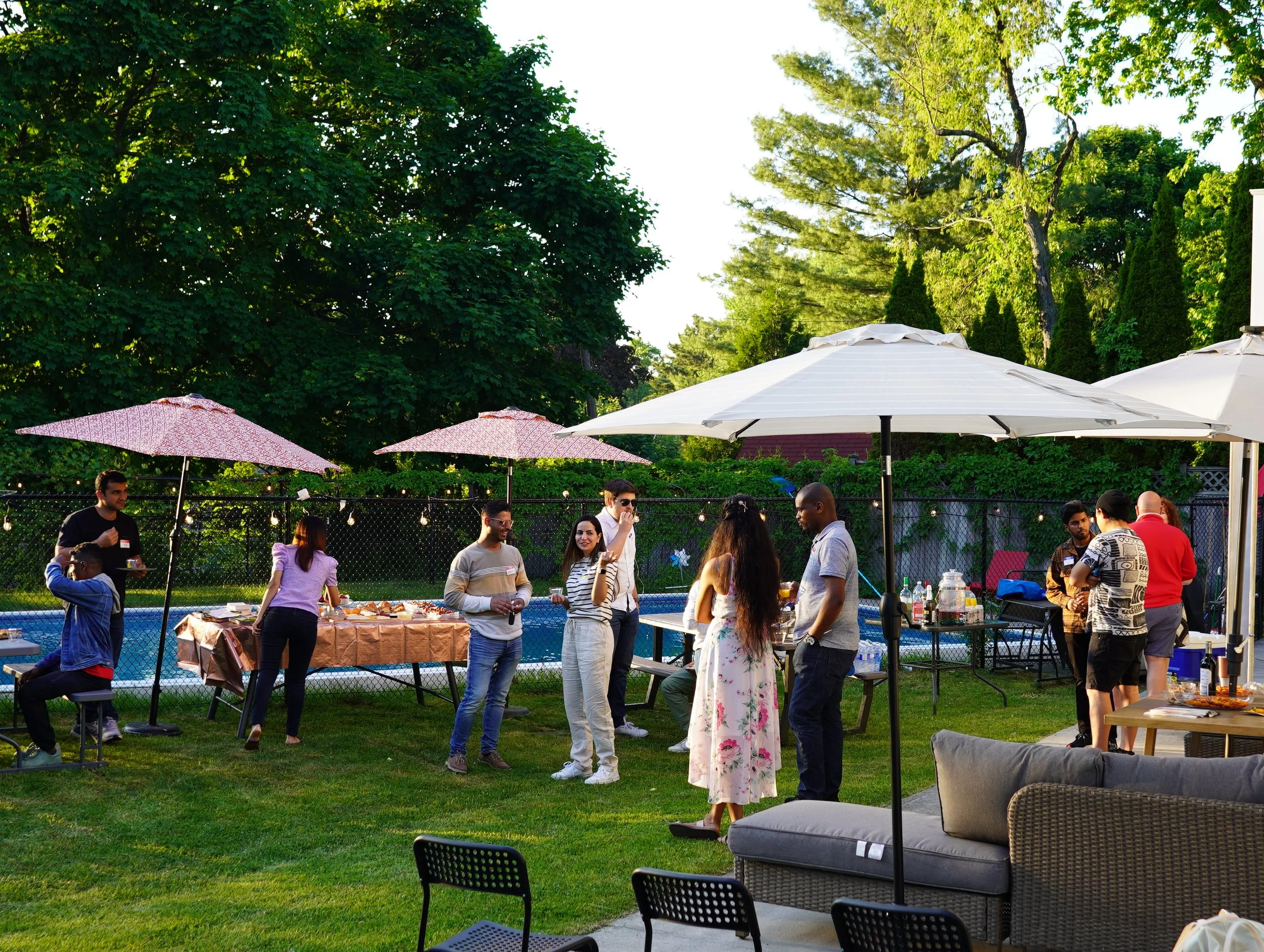 People enjoying a backyard party with food and drinks on a sunny day, surrounded by trees and umbrellas.