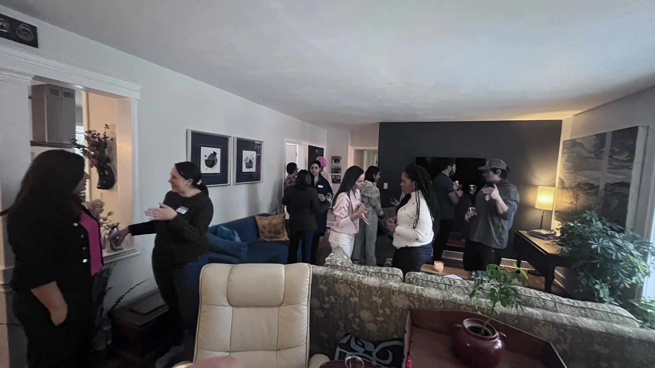 People gathered in a living room socializing, some holding drinks, with a dark accent wall, artwork, a TV, and a lit lamp in the background.