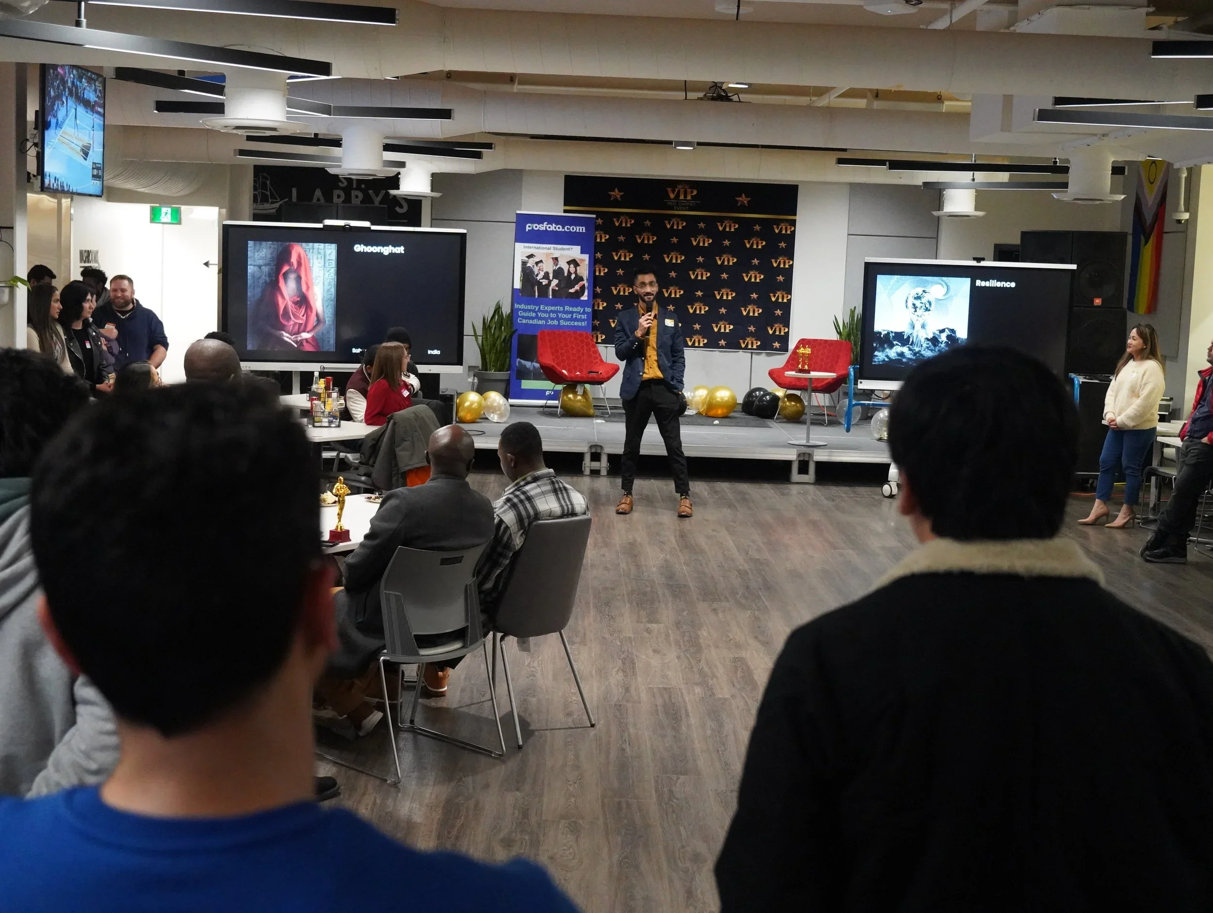 Business conference or presentation taking place in an indoor venue with a stage, audience seated at tables, and a speaker speaking into a microphone. Large screens display visuals behind the speaker, and there are VIP banners and balloons.