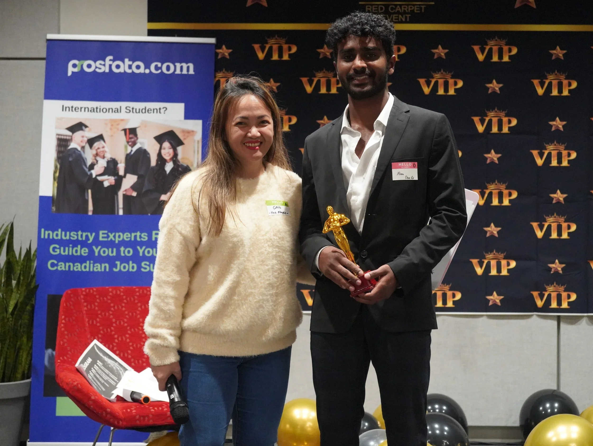 A woman and a man standing together on a stage at an award event. The woman is holding a microphone, and the man is holding a gold trophy. Behind them is a backdrop with 'VIP' and stars, and to their left is a banner with a picture of graduates and the website 'pros-fata.com'.