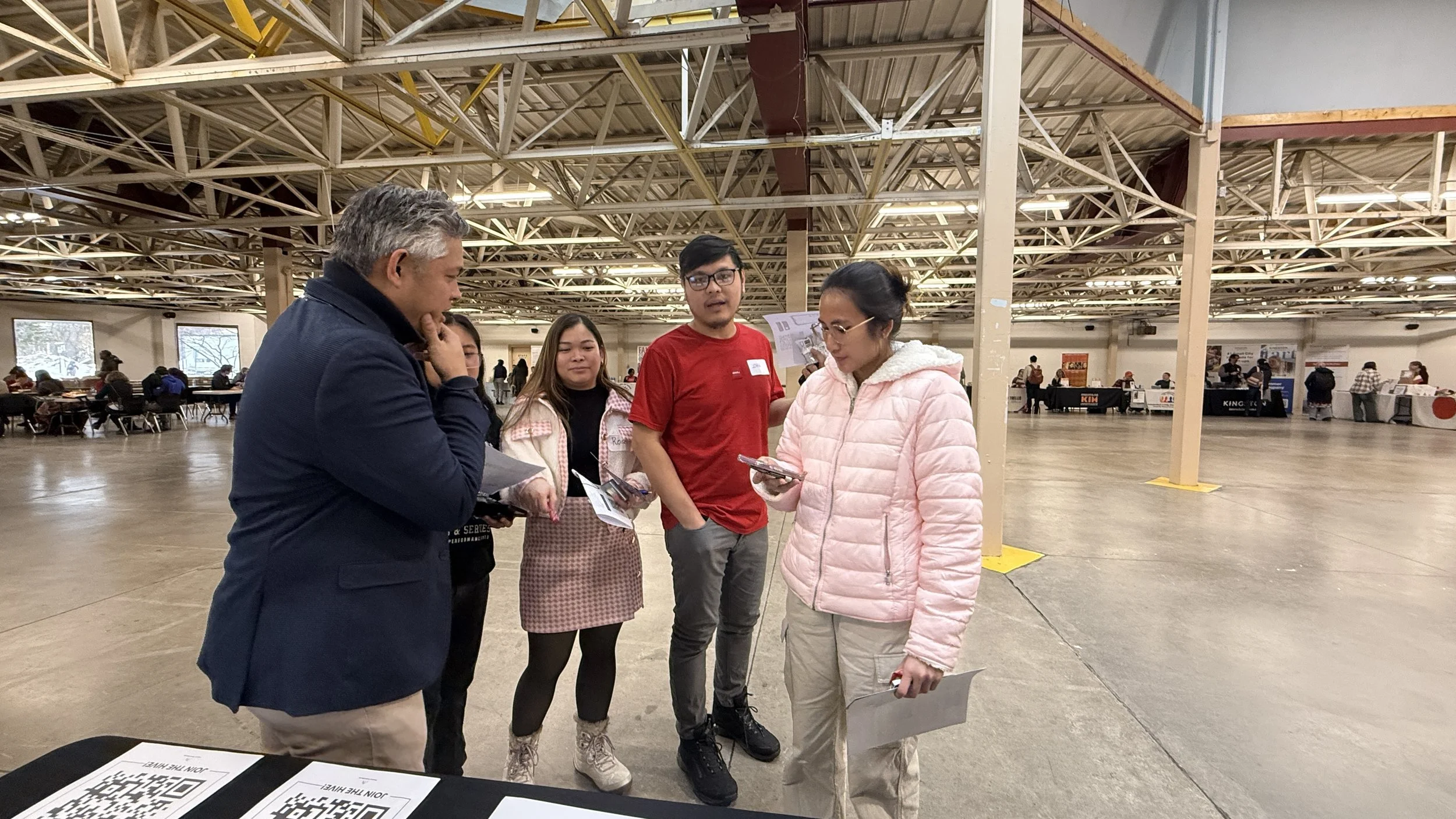 People standing and discussing in a large indoor event space, with tables and booths in the background.