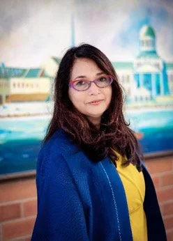 Woman with dark hair and pink glasses wearing a blue jacket and yellow shirt standing indoors with a painted cityscape background.
