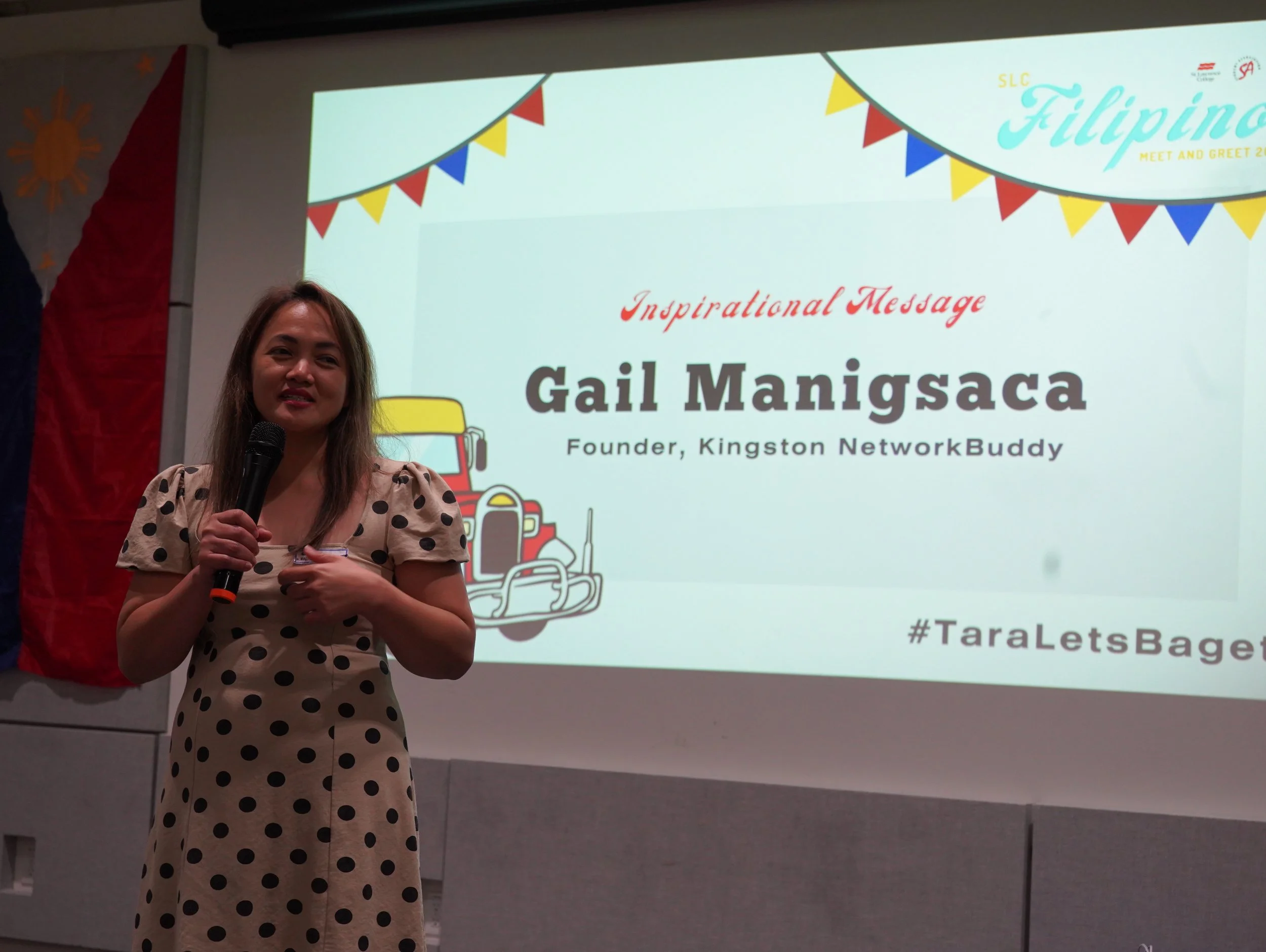 A woman wearing a polka dot dress holding a microphone speaks in front of a presentation slide that reads 'Inspirational Message, Gail Manigsaca, Founder, Kingston NetworkBuddy', with colorful bunting and a cartoon truck illustrated on the slide.