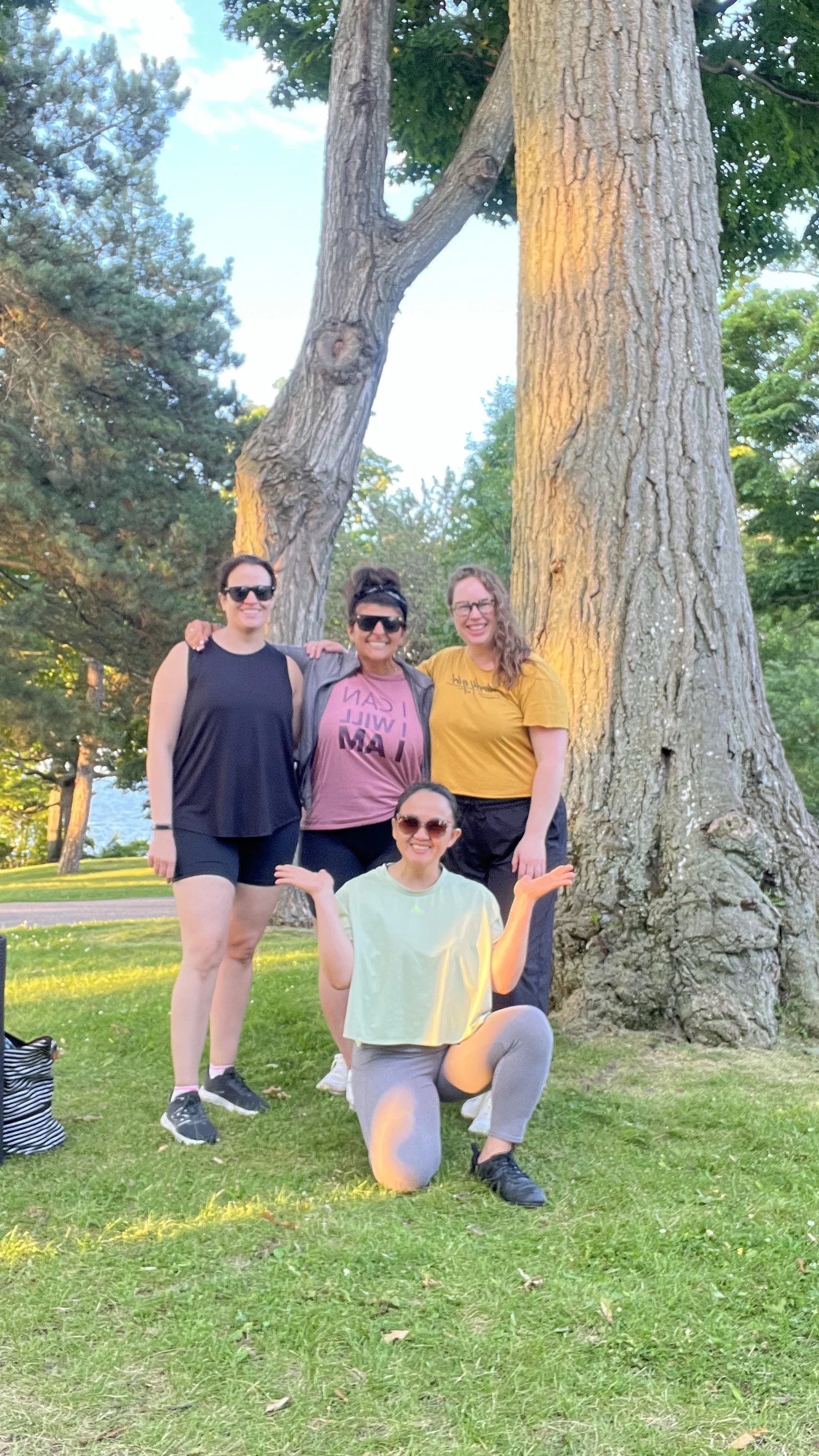 Four women are smiling and posing for a photo outdoors next to a large tree in a park, with grassy ground and trees in the background.