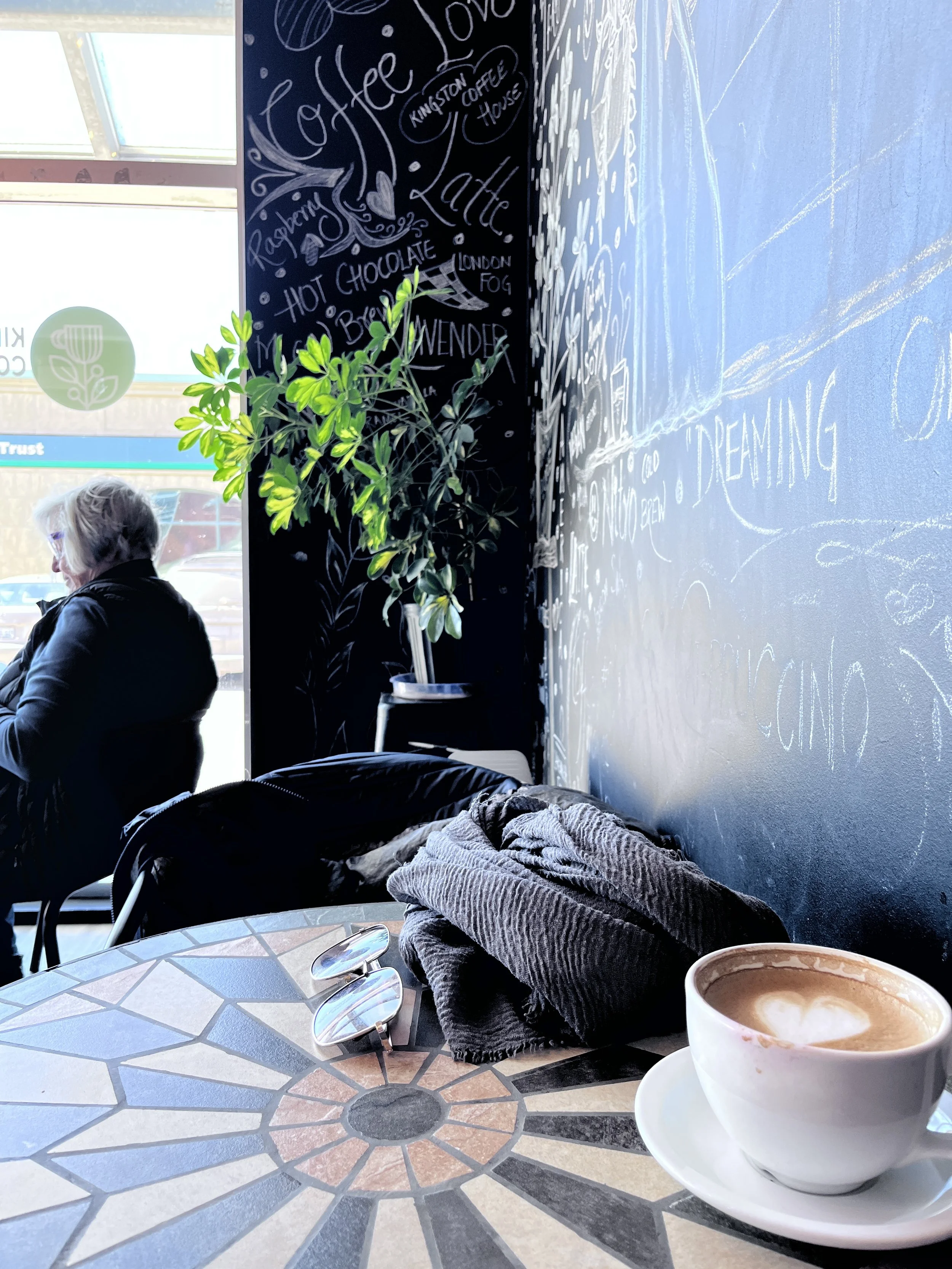 Inside of a coffee shop with a cup of coffee on a table, a folded gray sweater, and sunglasses. In the background, a woman with white hair is sitting near a large window. A black wall with white chalk writing and drawings about coffee is visible, along with a potted plant.