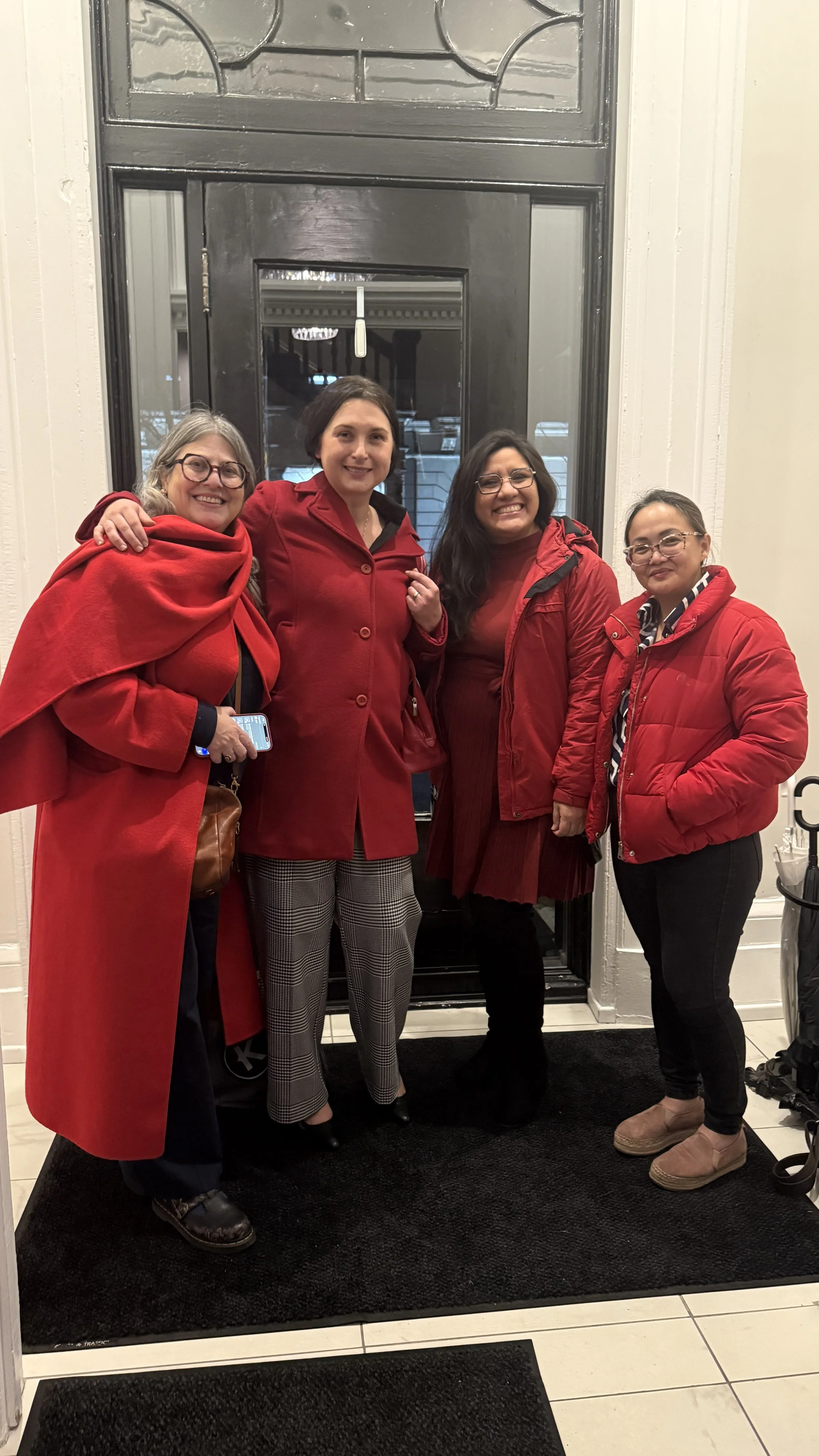 Four women dressed in red clothing standing in front of a black door, smiling, with a white wall and tiled floor in the background.