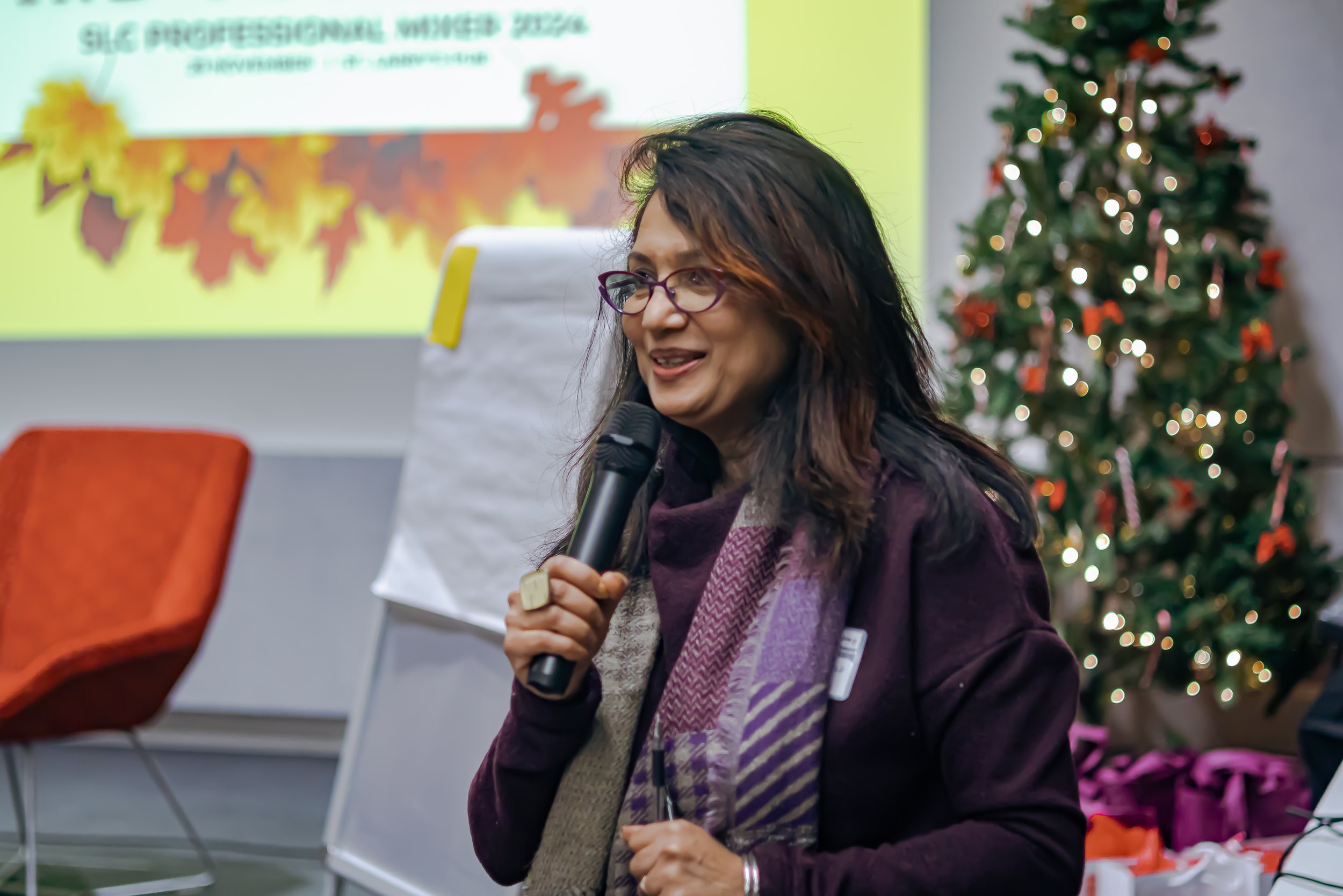 Woman speaking into a microphone at an indoor holiday event with a decorated Christmas tree in the background.