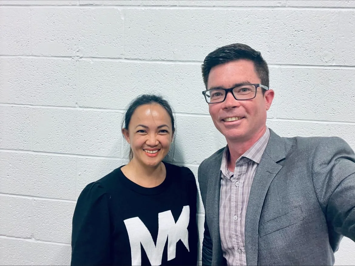 A woman and a man taking a selfie against a white brick wall. The woman wears a black shirt with the letter 'M' on it, and the man wears glasses, a gray blazer, and a plaid shirt.