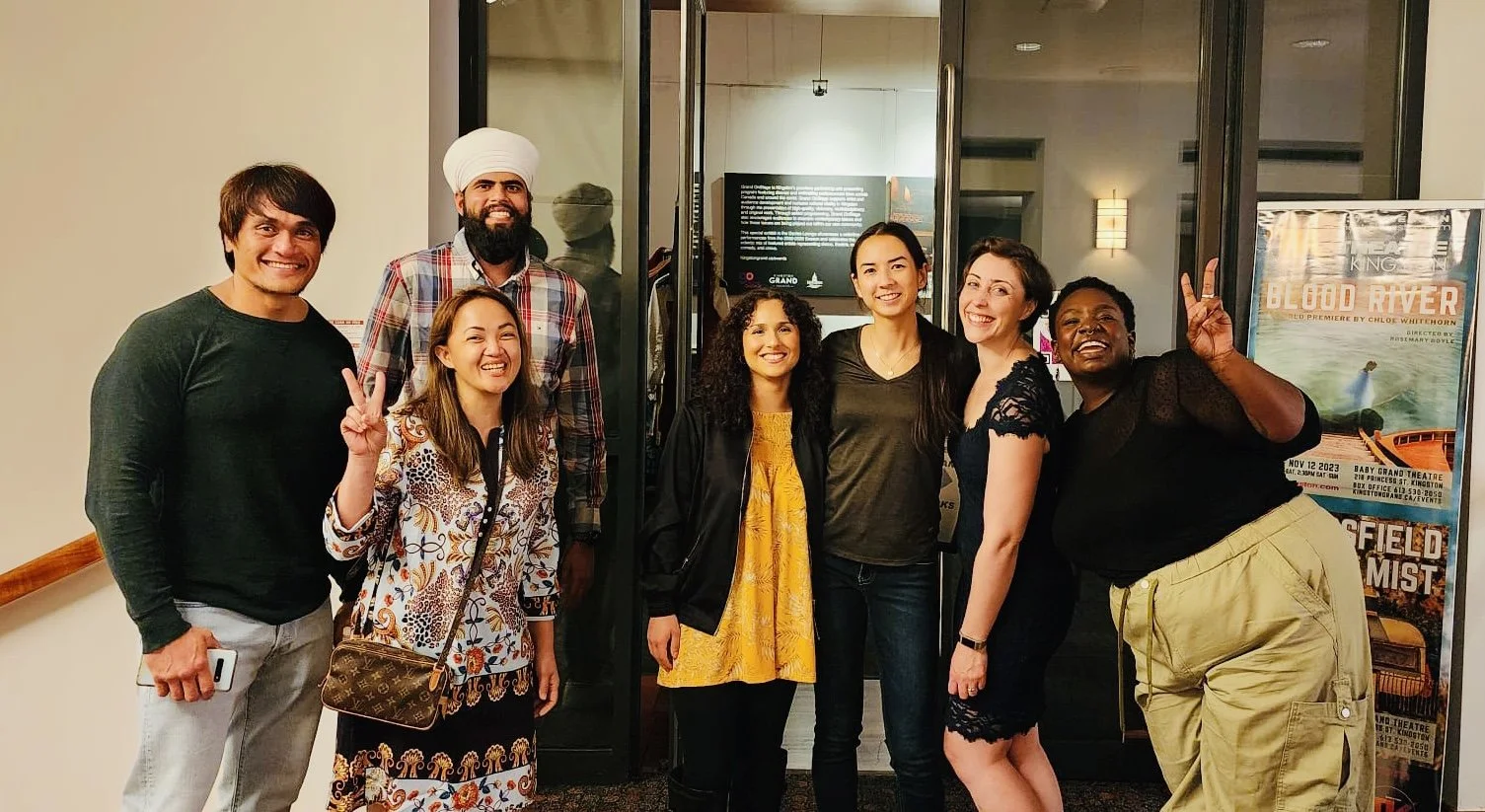 A group of eight people standing together inside, smiling and posing for a photo in front of a glass door with a poster to the right. The group includes men and women of diverse ethnicities, some making peace signs.