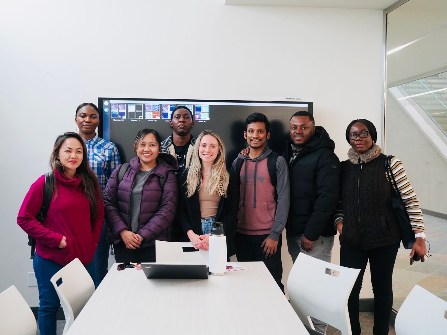 A group of nine diverse people standing together in a modern room, some smiling and others with neutral expressions, with a digital display behind them showing various images and icons.