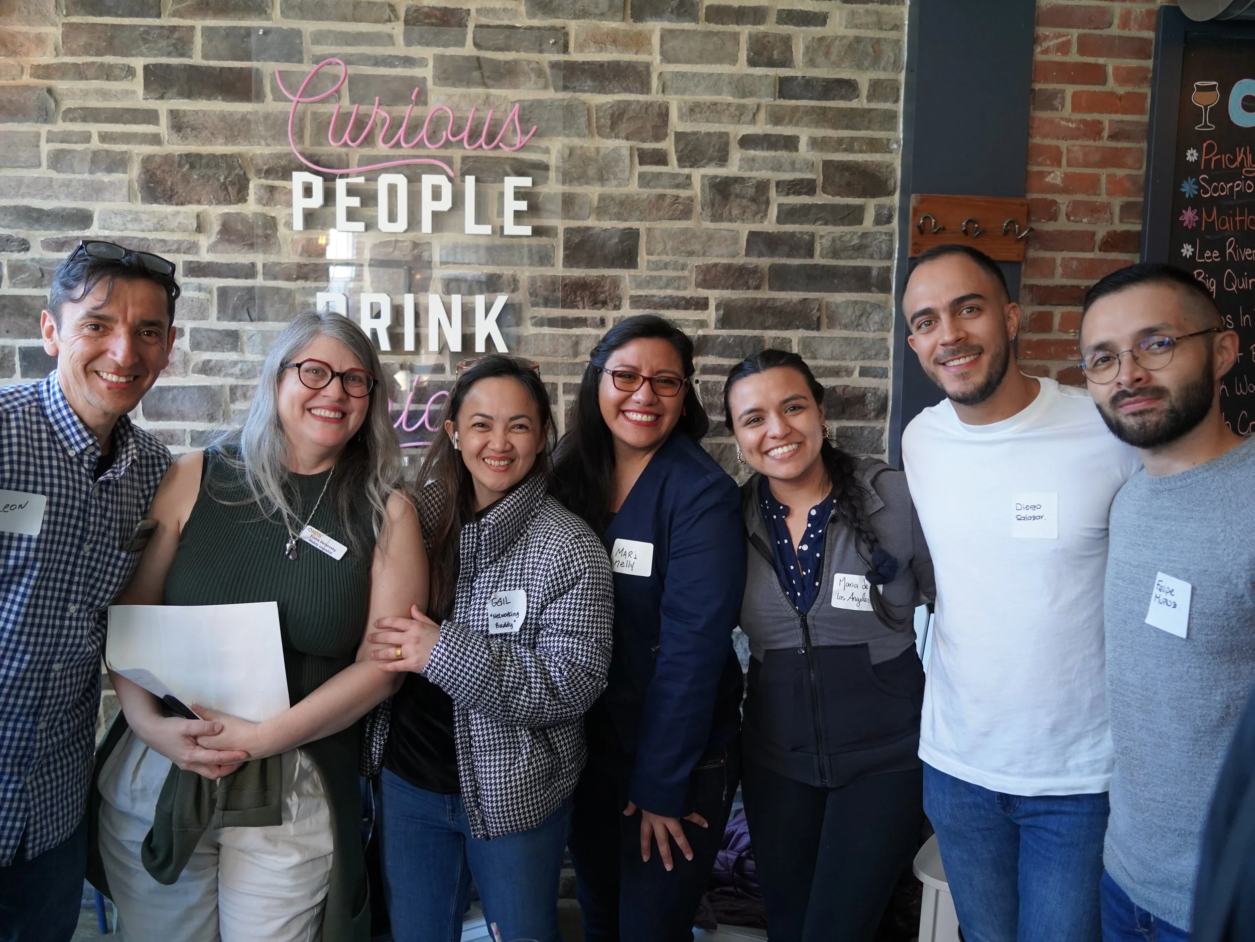A group of seven diverse adults standing together indoors, smiling at the camera, in front of a brick wall with a neon sign that says 'Curious People Drink'.