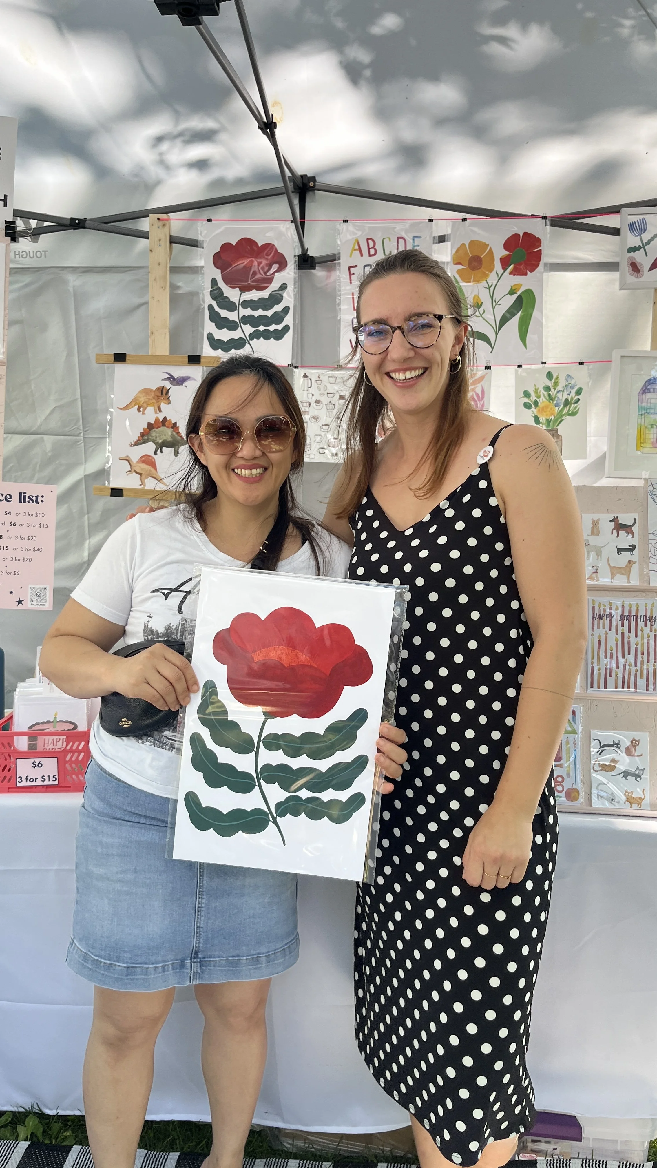 Two women smiling at an art booth, one holding a large printed art piece of a red flower with green leaves.
