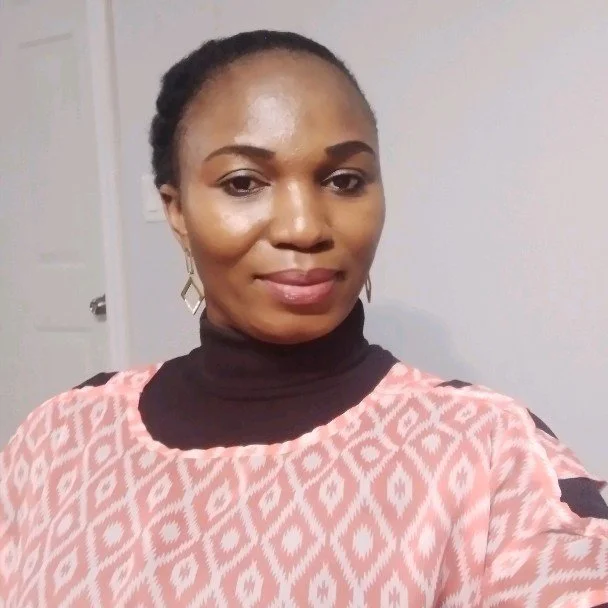 Portrait of a woman wearing a pink patterned top and a black turtleneck, with earrings, smiling in front of a grey wall.