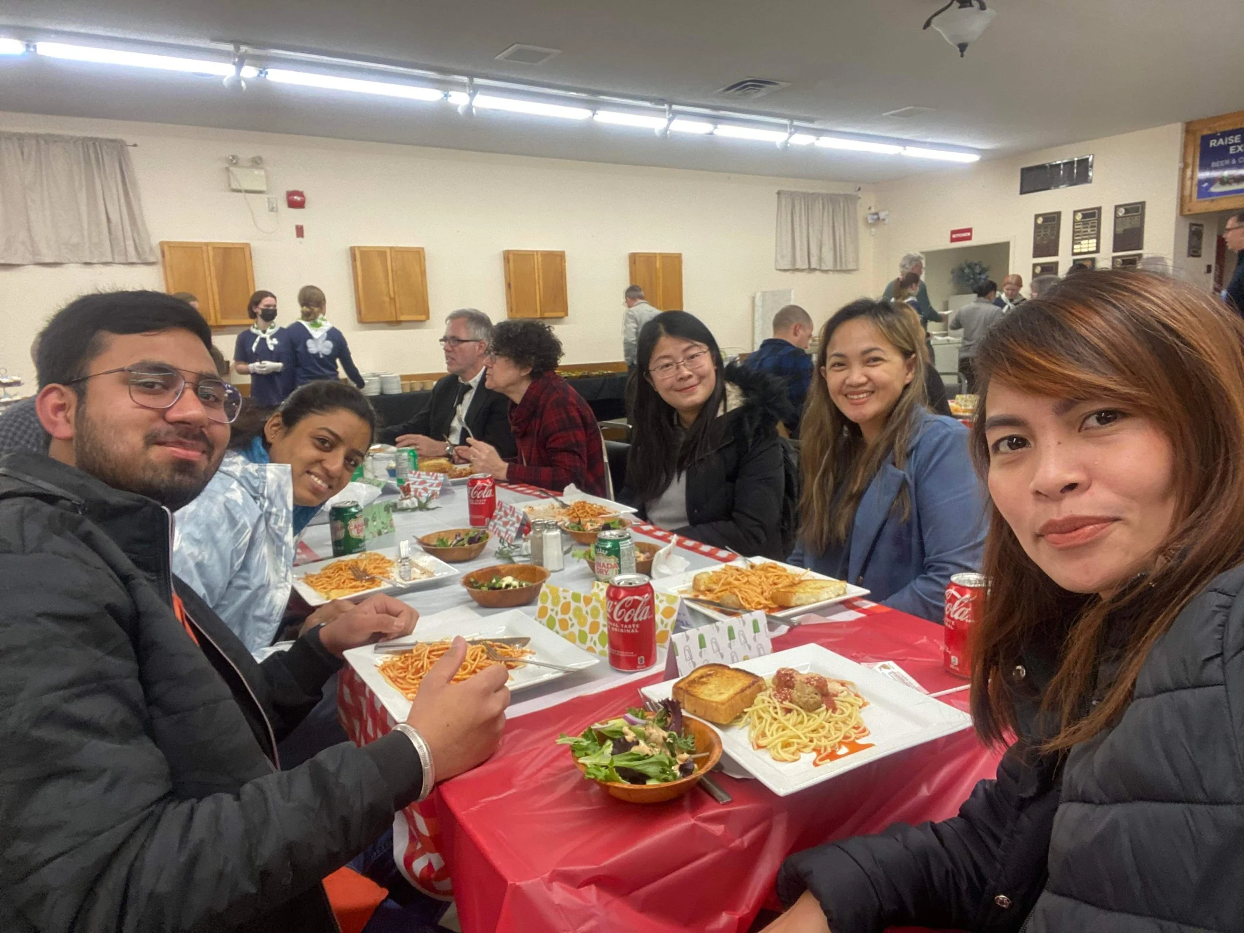 Group of six people sitting around a table at a festive meal, with plates of spaghetti, salads, bread, and cans of Coca-Cola, in a decorated indoor setting with other people dining in the background.