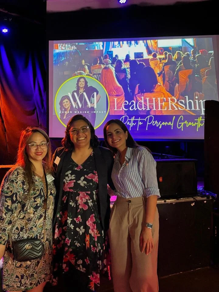 Three women standing together in front of a slide that reads "Women Making Impact" and "Leadership: Path to Personal Growth". They are smiling and dressed in business casual attire, with a dark stage curtain and colorful lighting in the background.