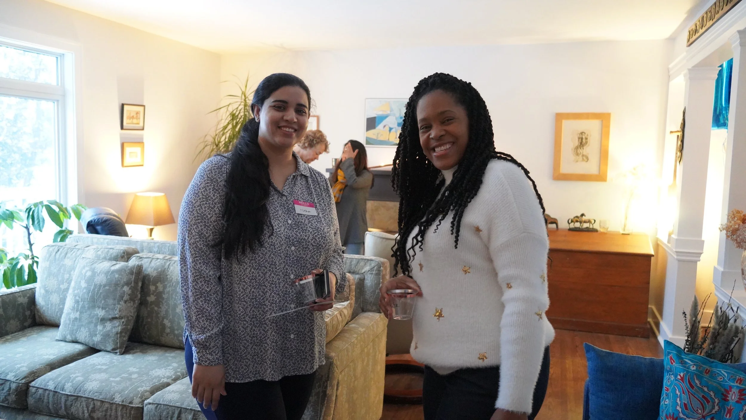 Two women smiling indoors, one wearing a patterned blouse and a name tag, holding a drink, the other in a white sweater with stars, also holding a drink, with a cozy living room background.