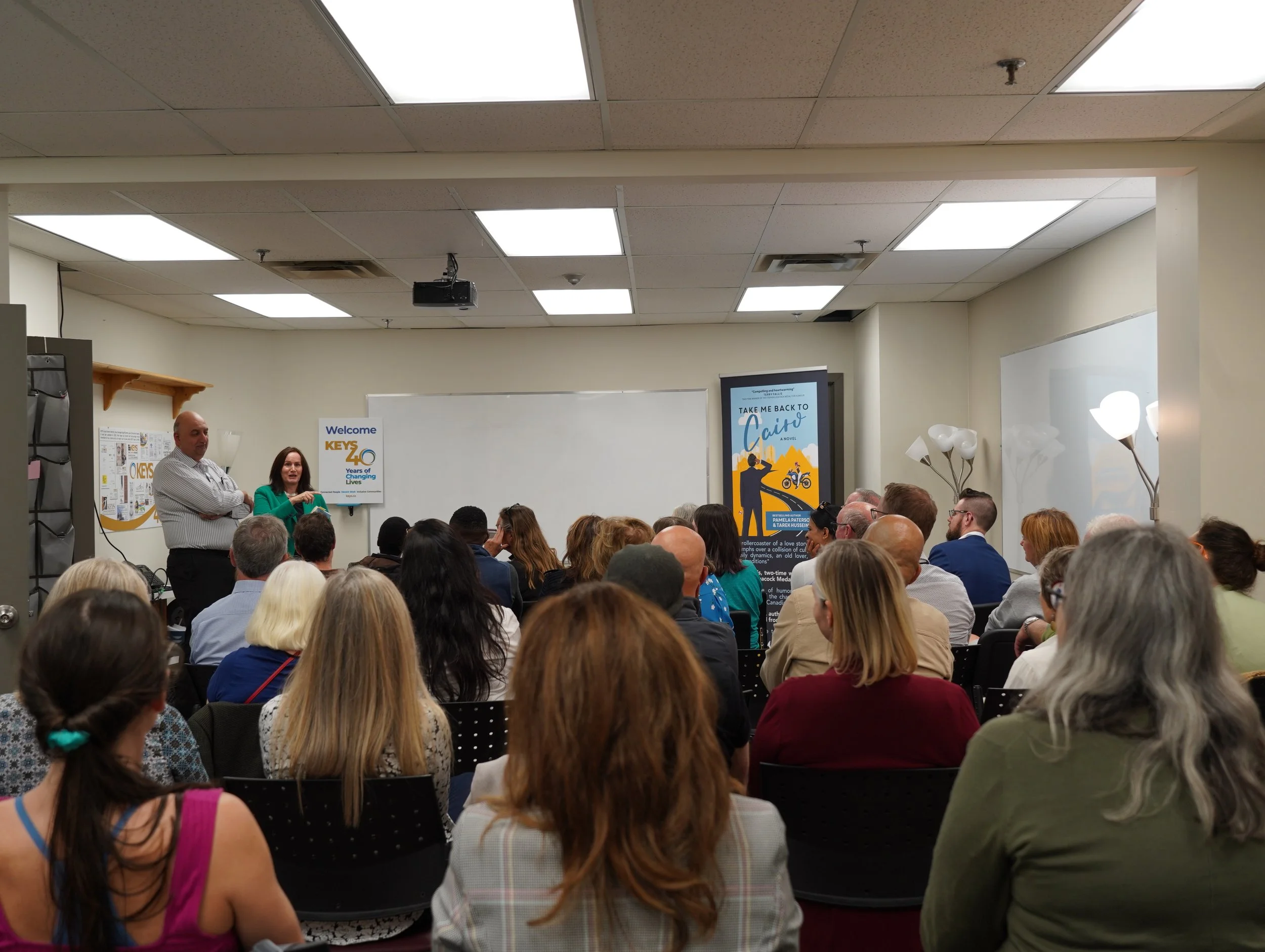 A room filled with people attending a presentation or conference, with two presenters at the front near a whiteboard and digital display.