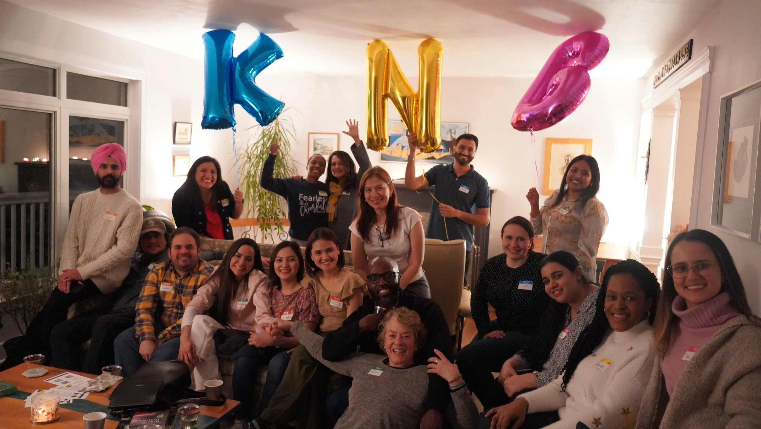 Group of people celebrating with balloons in a cozy living room, some holding large letter balloons spelling "KIN" and pink balloon, with smiles and cheerful expressions.