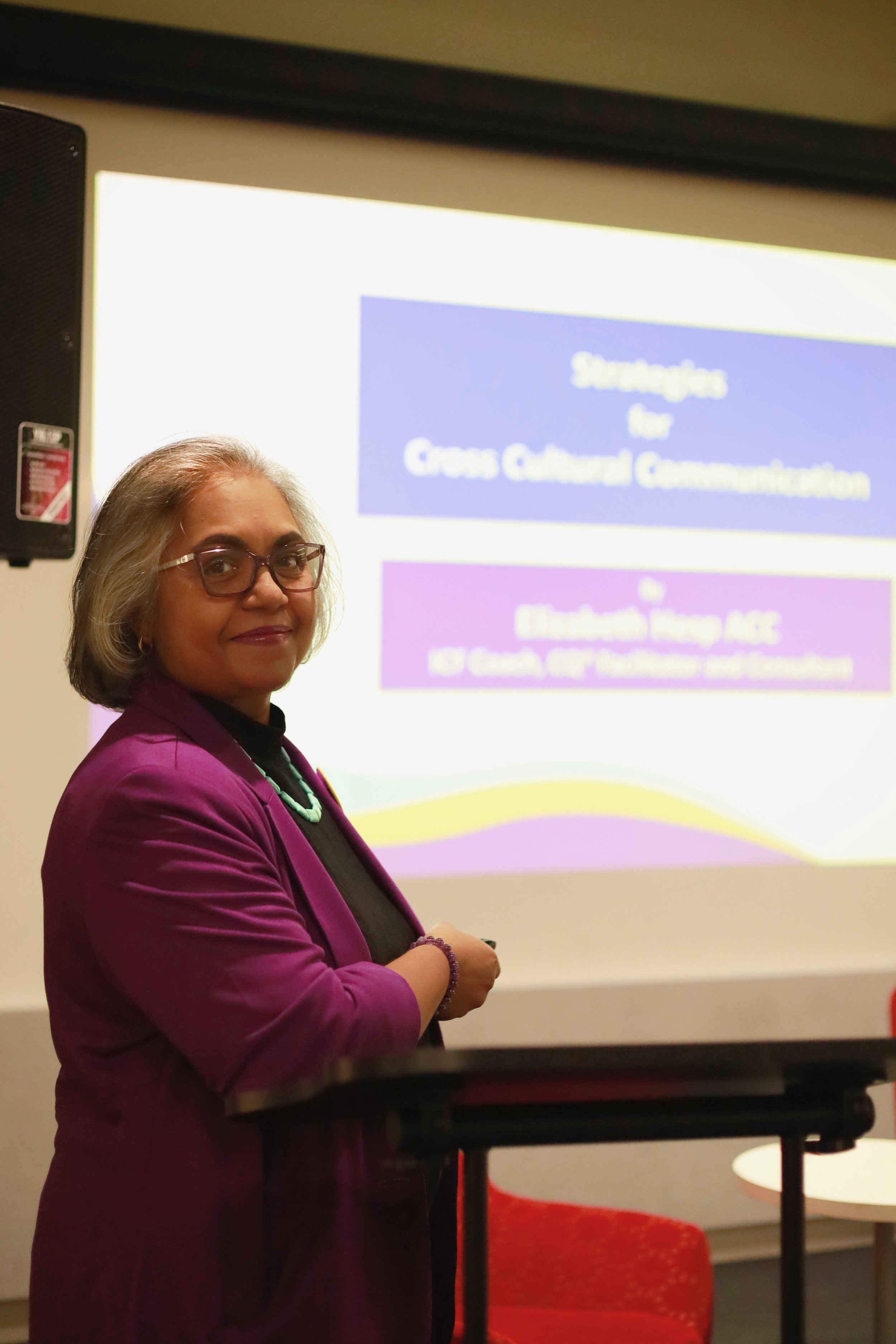 A woman with glasses and short gray hair wearing a purple blazer standing in front of a presentation slide about cross-cultural communication.