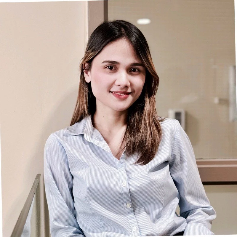 A young woman with long brown hair, wearing a light blue button-up shirt, sitting indoors against a beige wall, smiling at the camera.