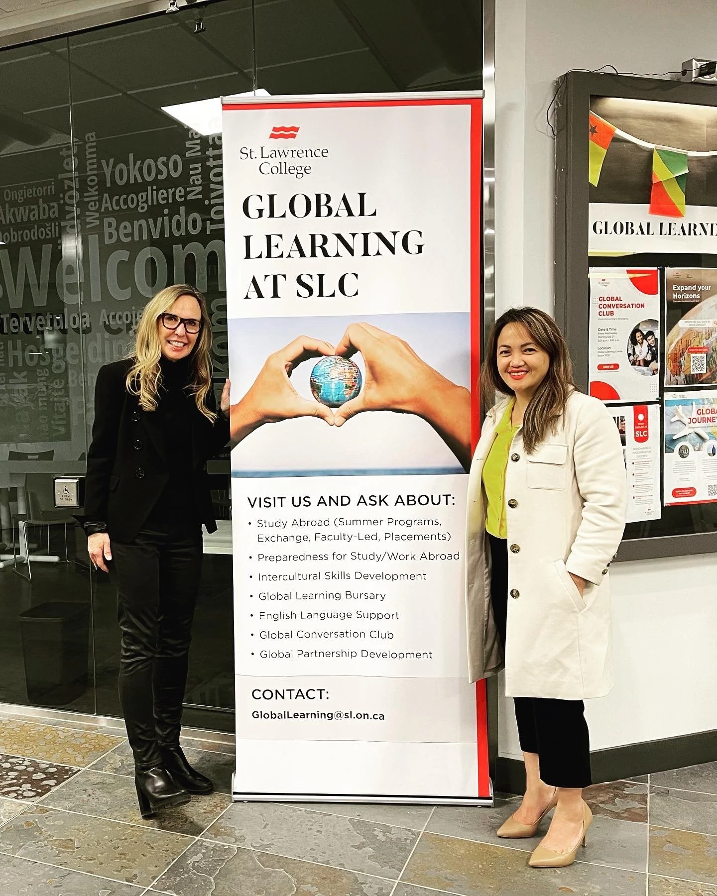 Two women standing next to a large vertical banner for St. Lawrence College, promoting global learning programs including study abroad, intercultural skills, and international support. They are inside a building with glass doors and informational posters visible behind them.