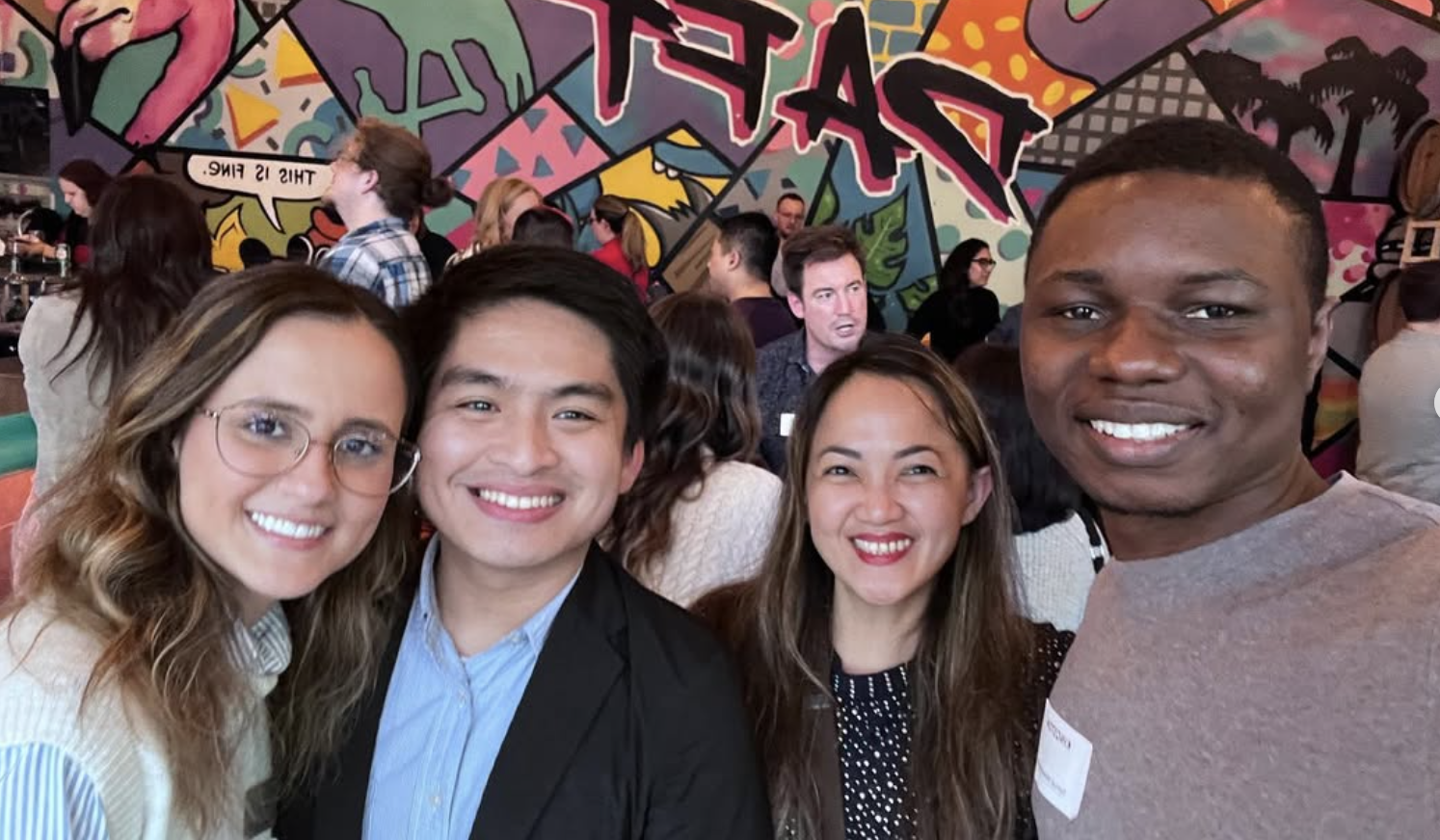 Four smiling people taking a selfie at a crowded indoor event with colorful graffiti art on the wall behind them.