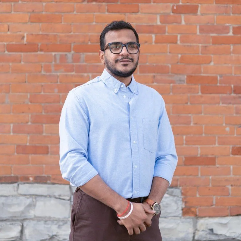 A man with glasses and a beard standing in front of a brick wall, wearing a light blue button-up shirt and brown pants.