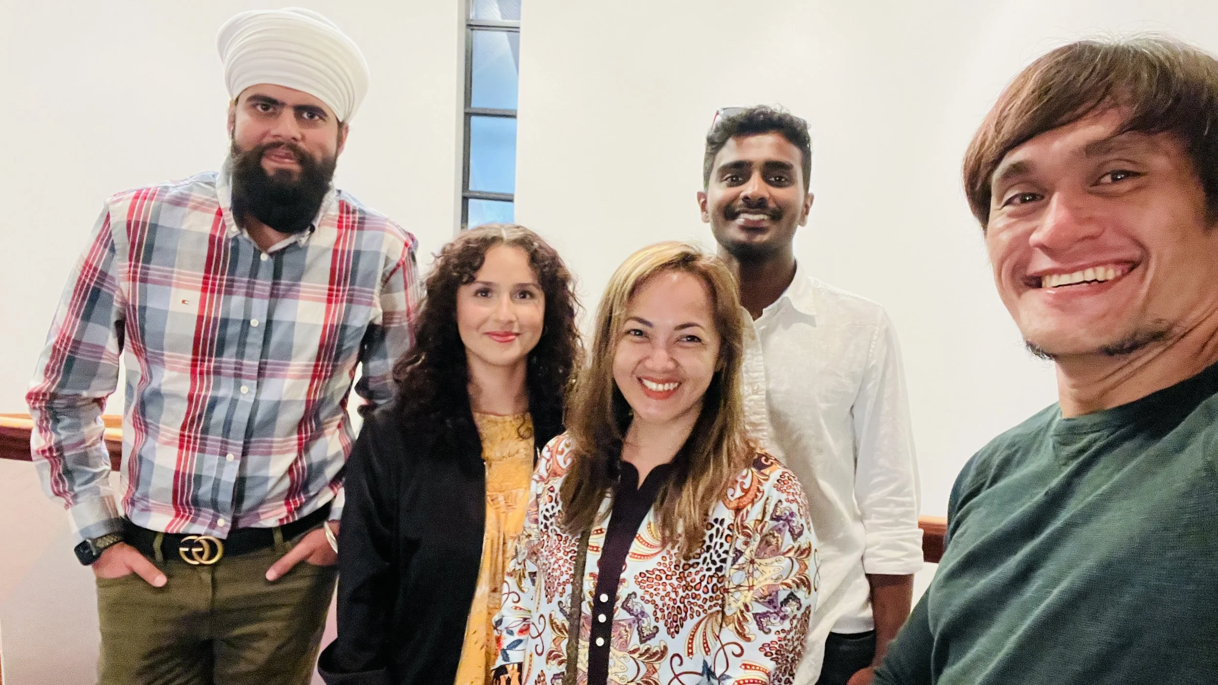 Five diverse individuals smiling and posing for a group photo indoors, with a plain white wall and a window in the background.
